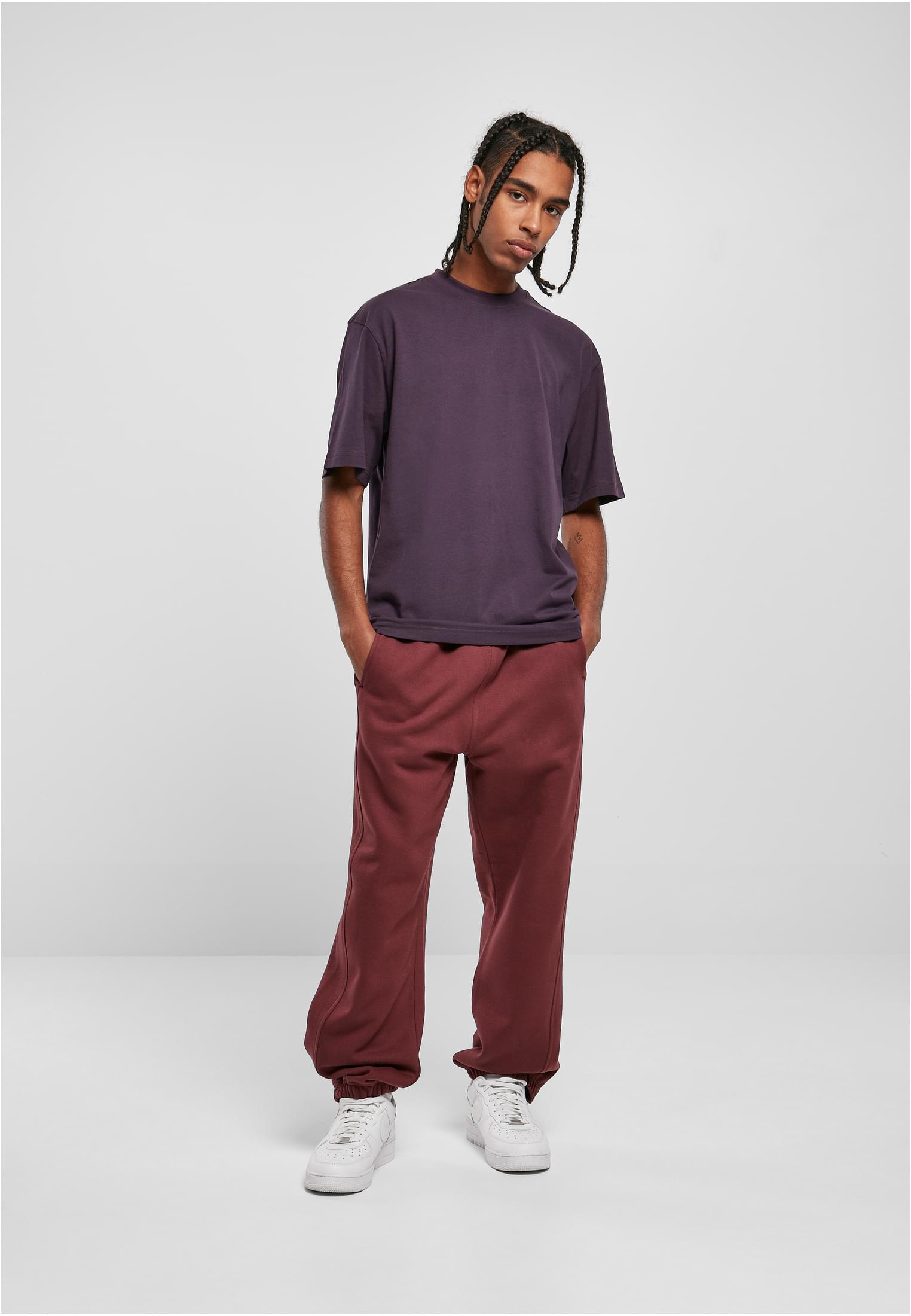 Manlig modell i helfigur som bär en mörklila Tall Tee, vinröda sweatpants och vita skor mot en ljusgrå bakgrund.