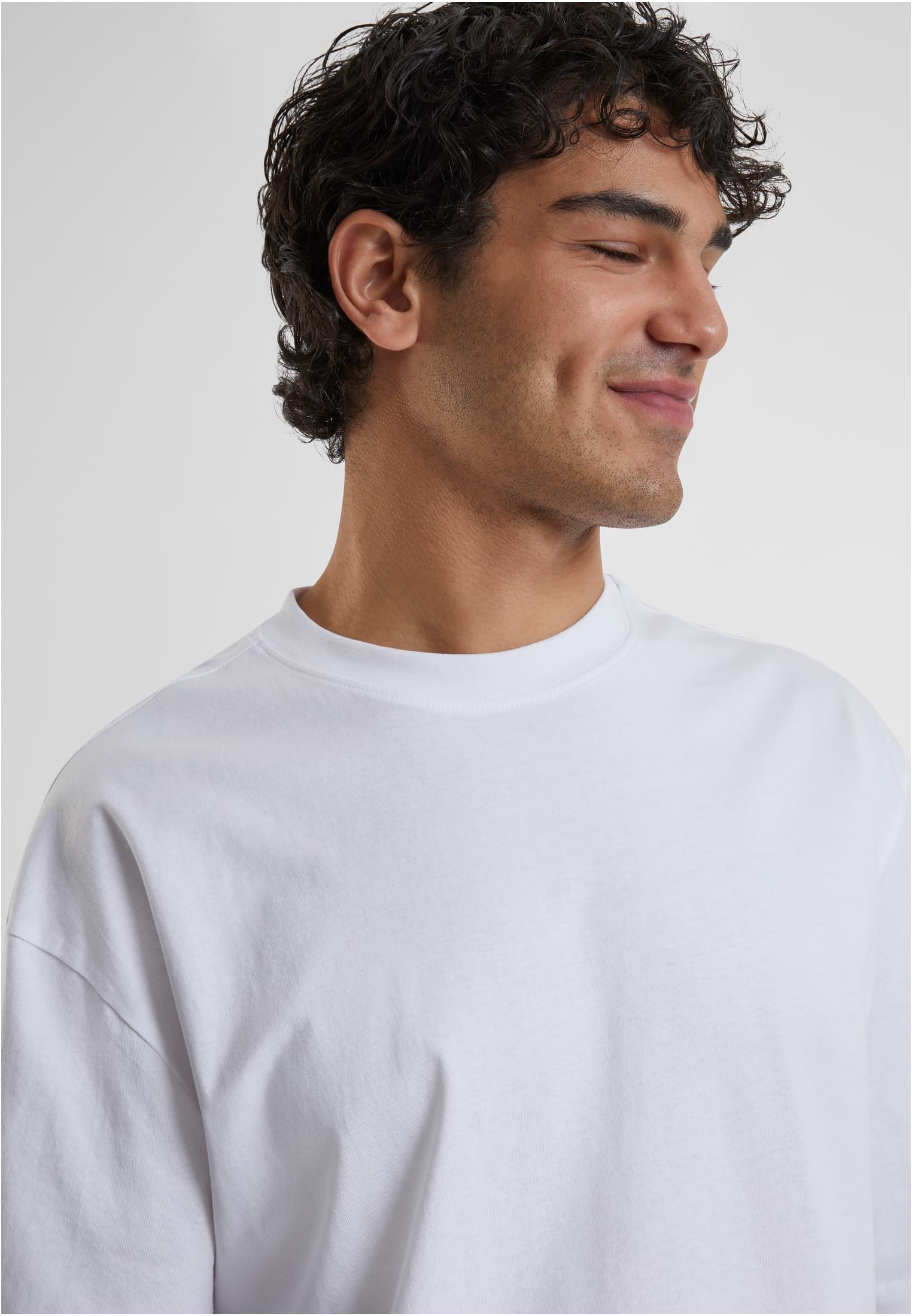 Närbild på en man med mörkt lockigt hår som ler med stängda ögon, iklädd en ljus Tall Tee.