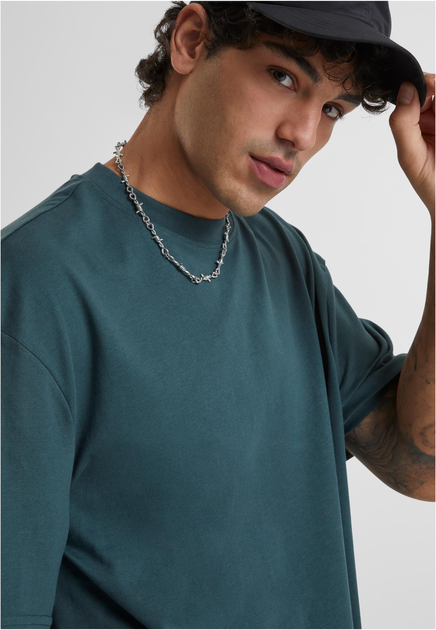 Närbild på en man som bär en mörkgrön Tall Tee t-shirt, en svart keps och ett taggtrådshalsband.