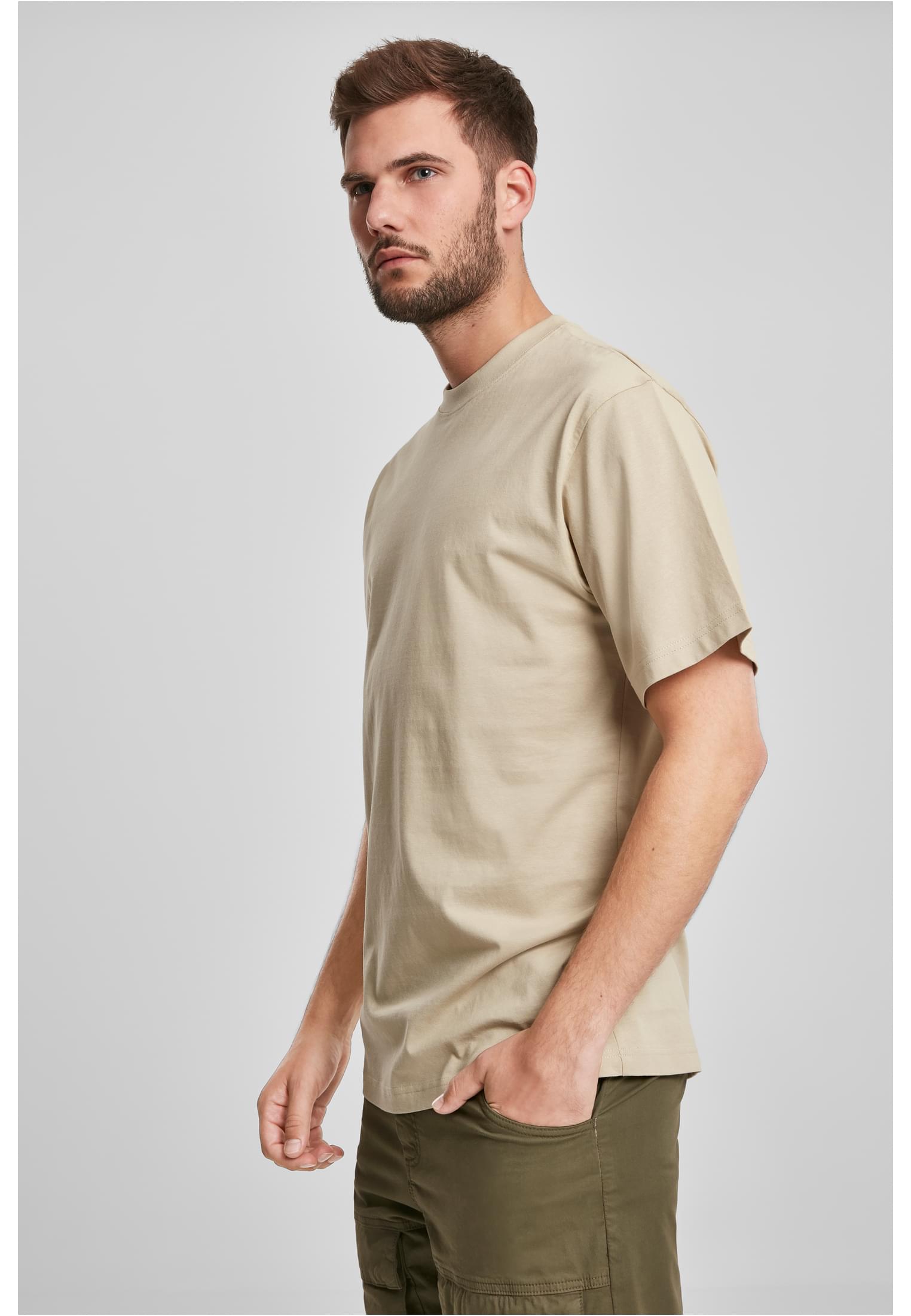 Manlig modell bär en beigefärgad Tall Tee och gröna byxor mot en ljusgrå bakgrund.