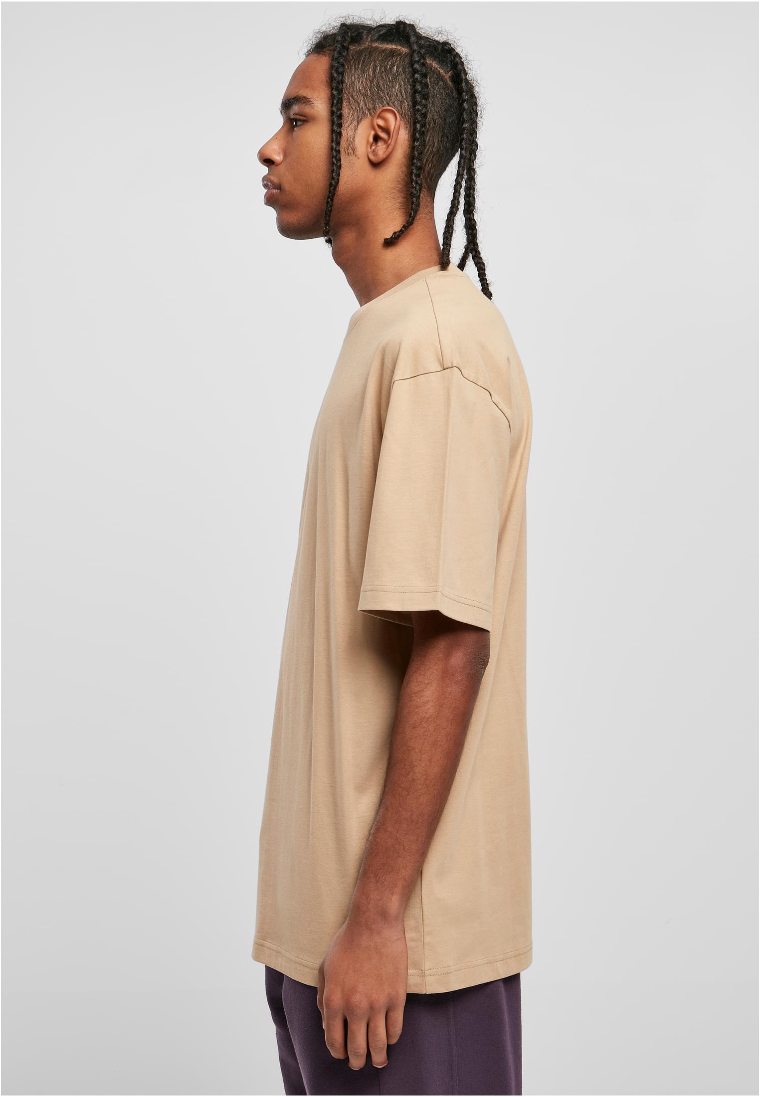En man med flätat hår, fotograferad från sidan, klädd i en beigefärgad Tall Tee med extra lång passform.