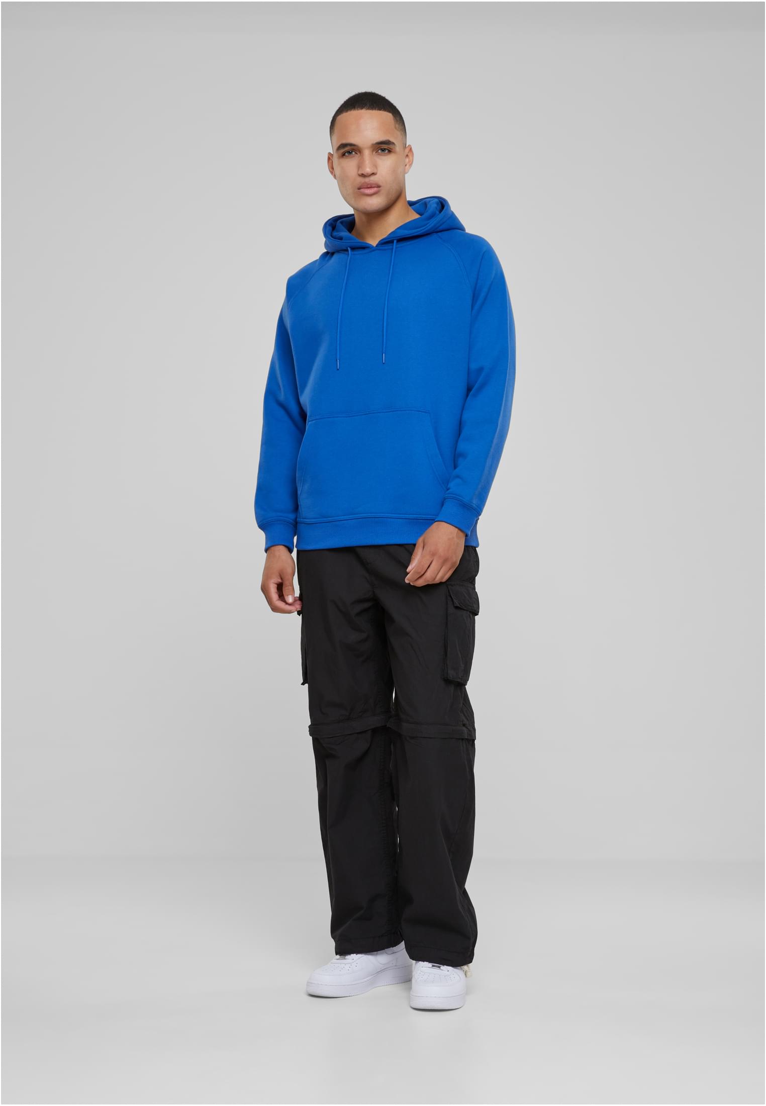 En man som bär en blå luvtröja, svarta cargobyxor och vita sneakers mot en ljusgrå bakgrund.