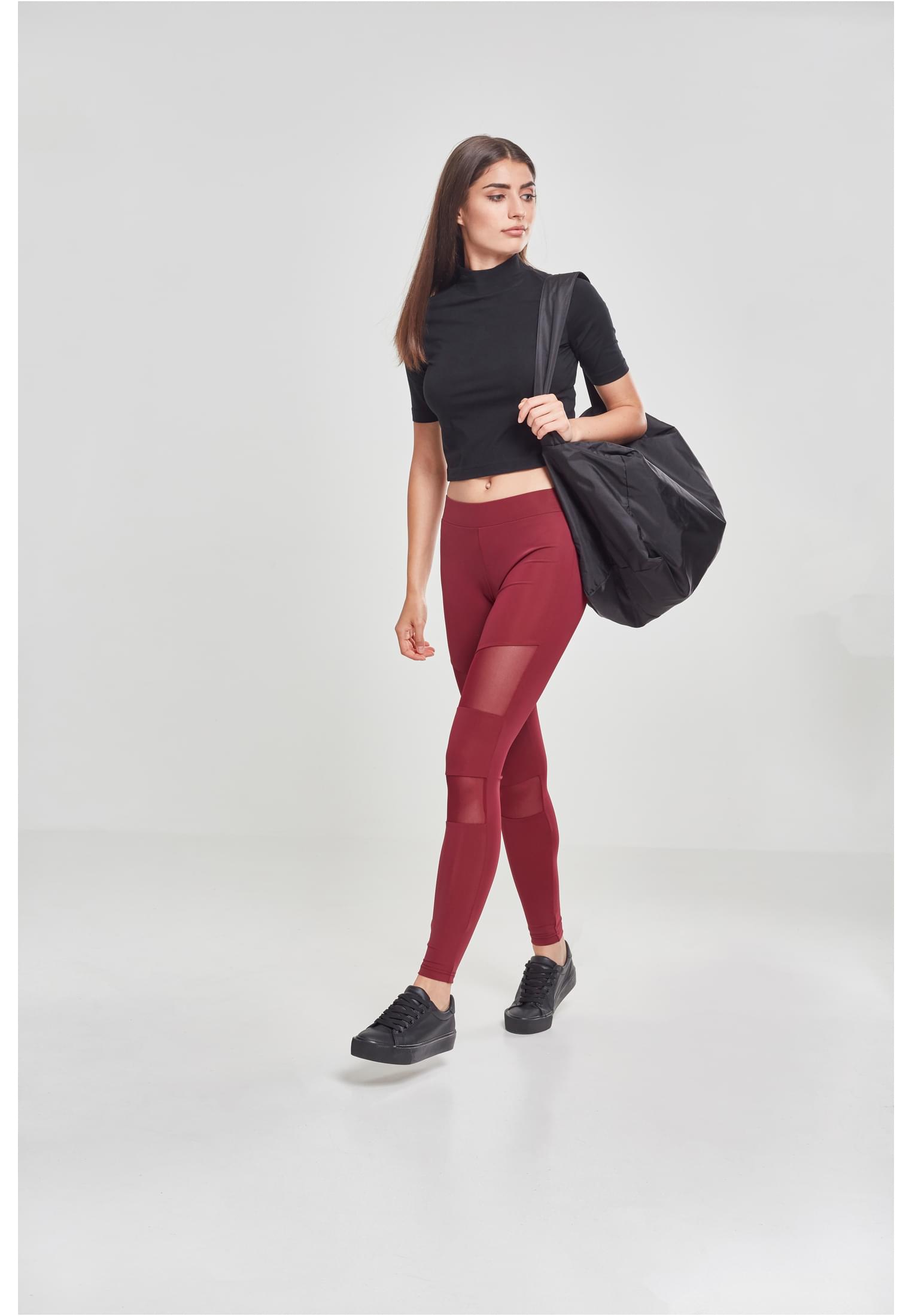 En kvinna går medan hon bär ett par vinröda Ladies Tech Mesh Leggings med transparenta nätpaneler, en svart topp och en stor svart träningsväska.