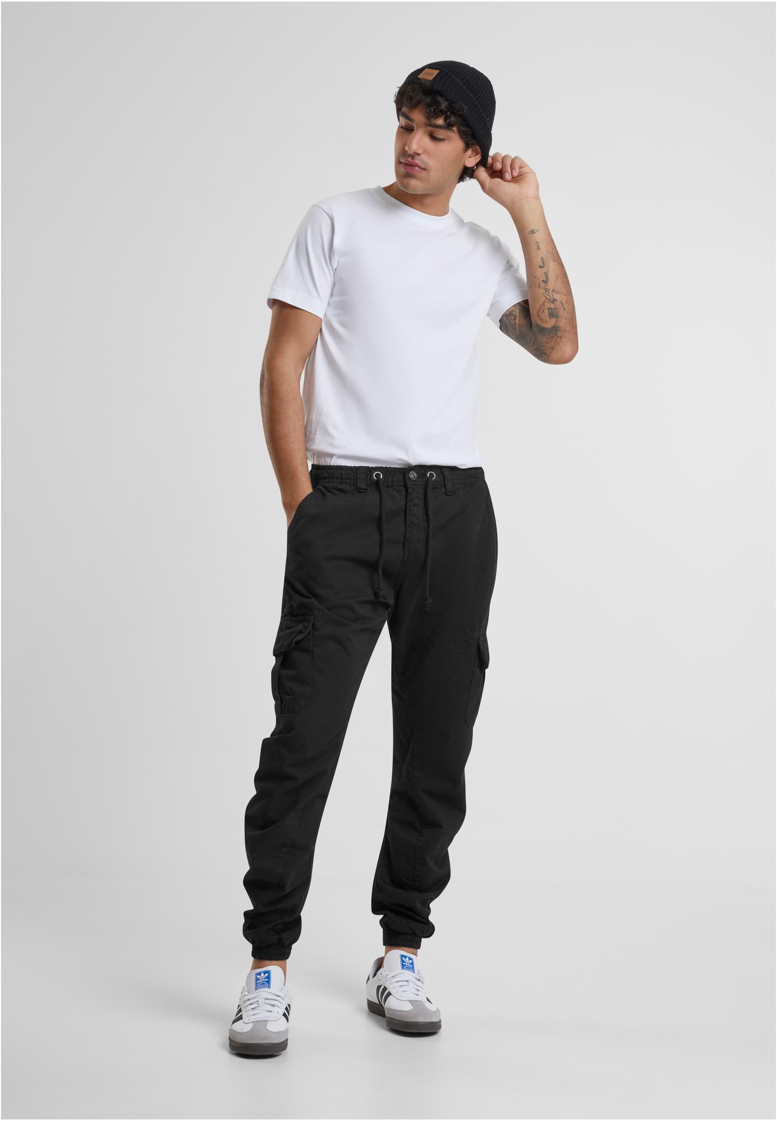 En manlig modell som bär svarta Cargo Jogging Pants, en vit T-shirt och en svart mössa.