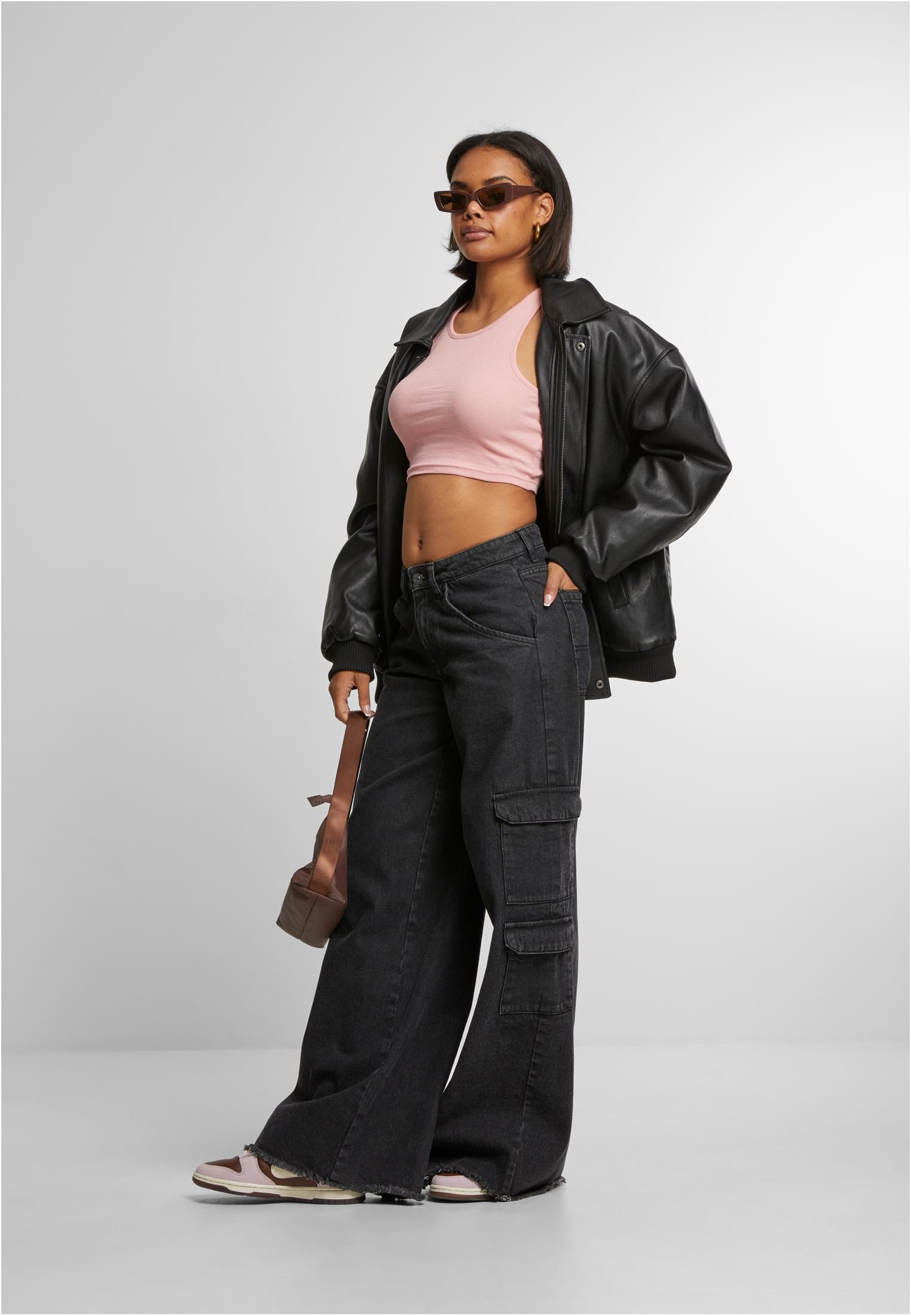 Helhetsbild av en kvinna som bär en ljusrosa croppad ribbad topp, stylad med en öppen svart skinnjacka, vida svarta jeans och solglasögon.