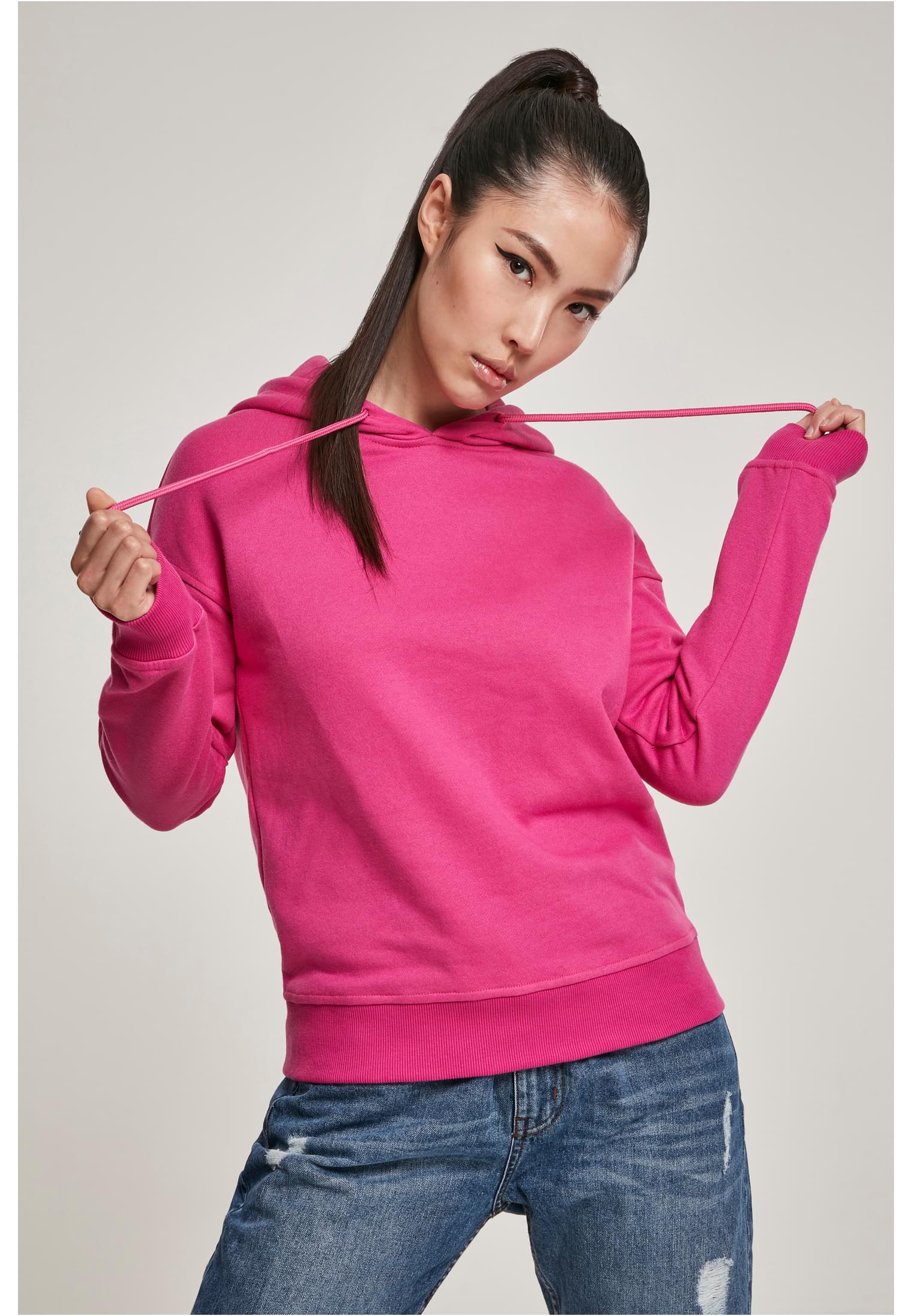 En kvinna med mörkt hår i hästsvans bär en rosa Ladies Hoody från Urban Classics och blå jeans.