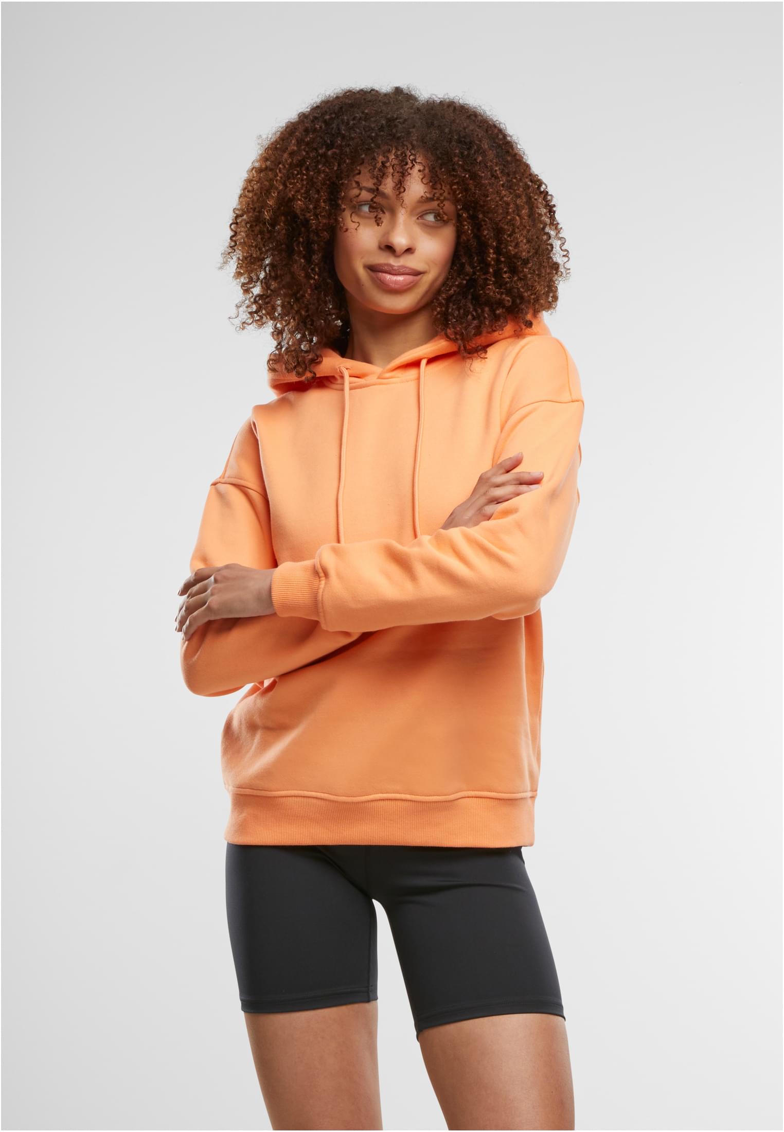 En kvinna poserar med armarna i kors i en ljusorange Ladies Hoody från Urban Classics och svarta cykelbyxor.