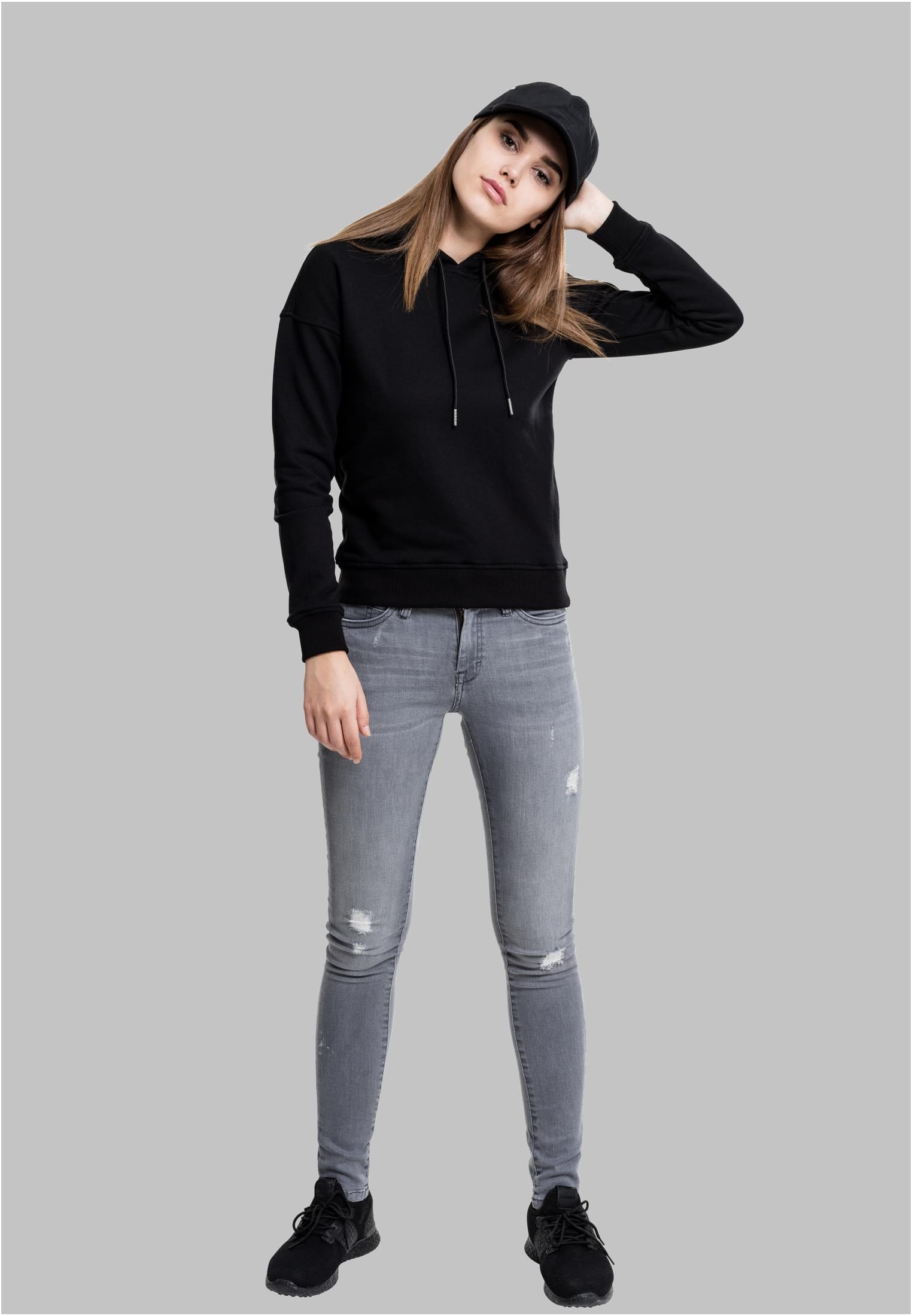 Helkroppsbild på en kvinna som bär en svart luvtröja, grå slitna jeans och en svart keps.
