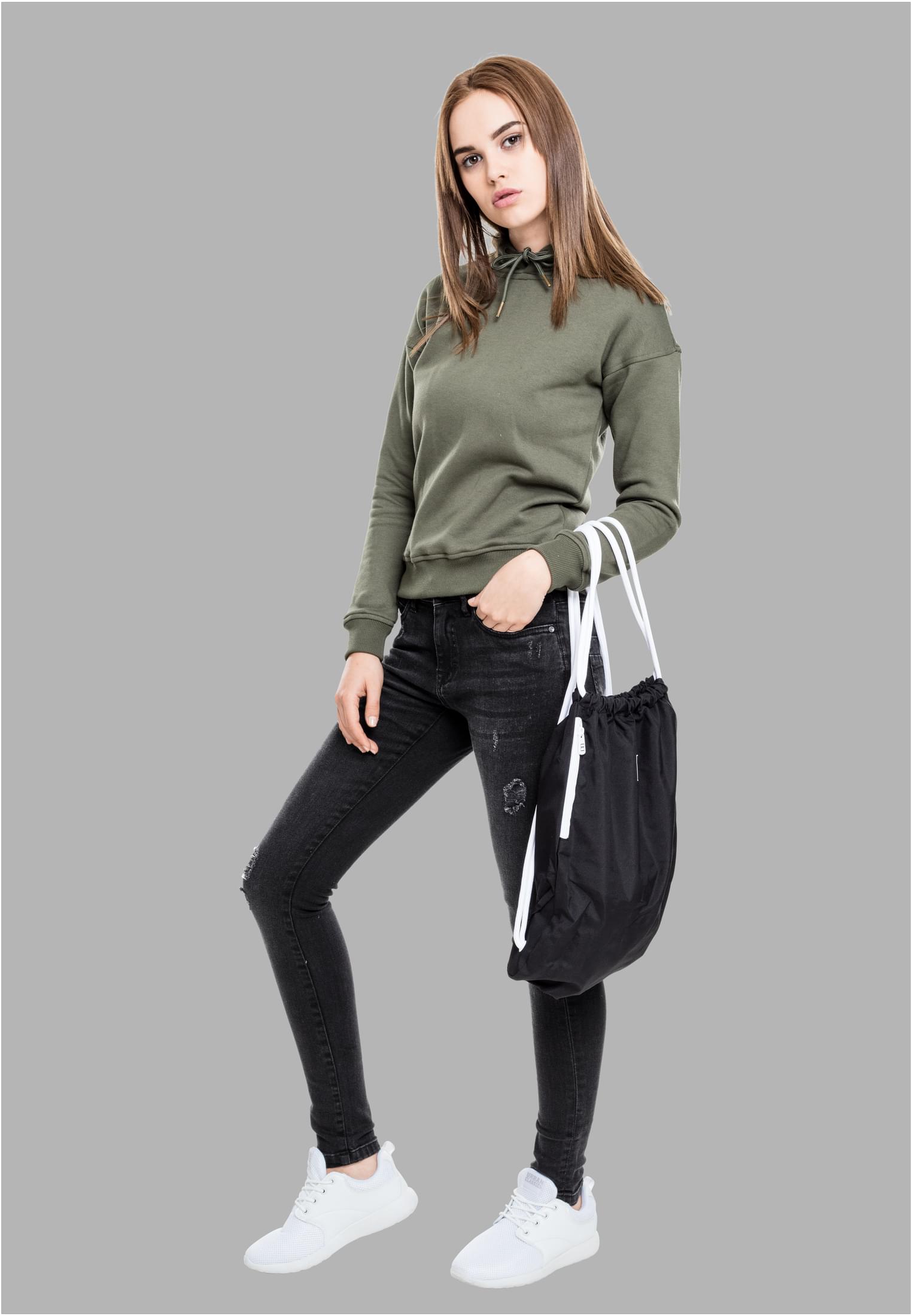 En kvinna som bär en grön luvtröja, svarta slitna jeans och vita sneakers. Hon står vänd framåt och bär en svart gympapåse på armen.