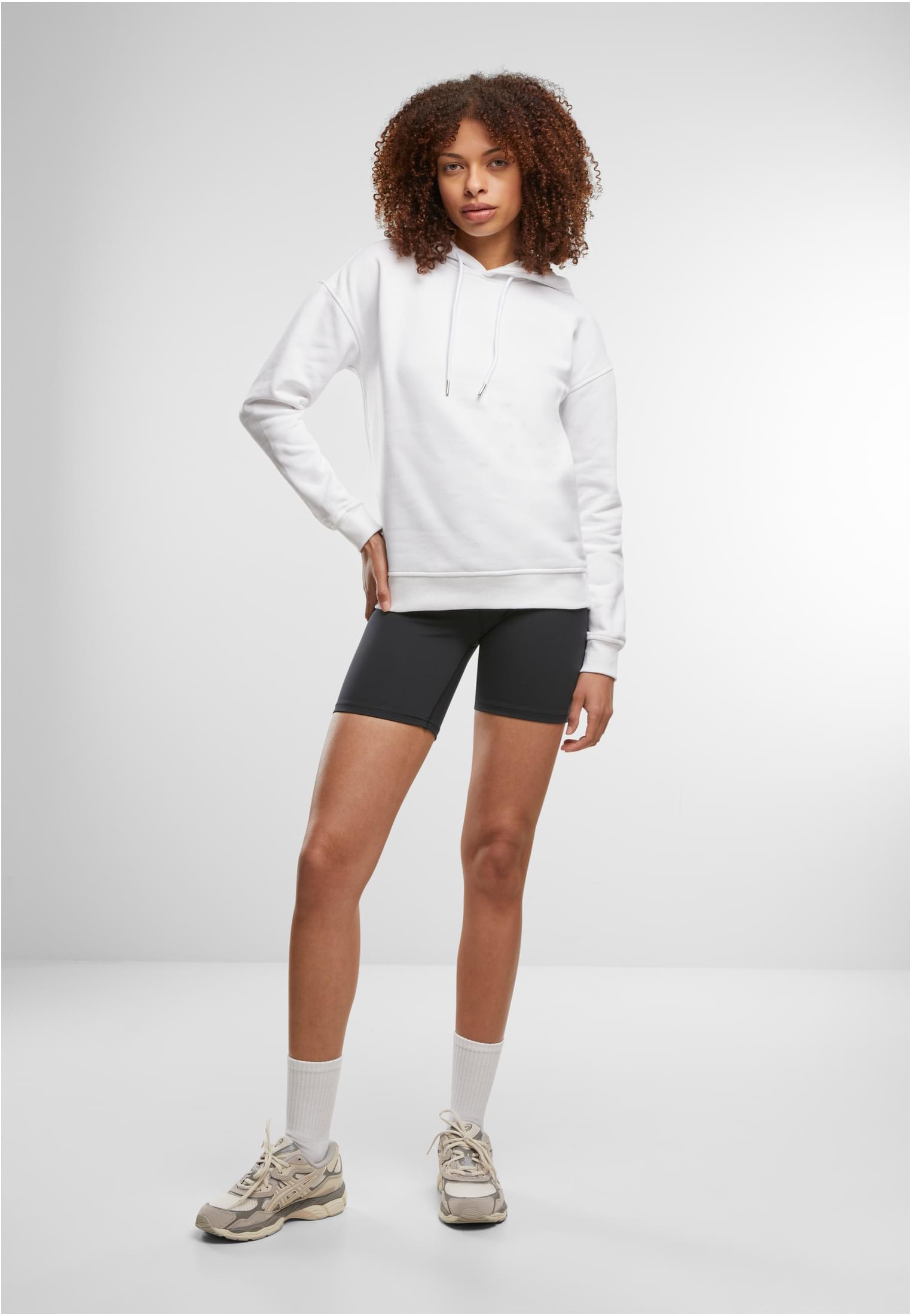 En modell i helfigur som bär en vit luvtröja, svarta cykelbyxor och sneakers.