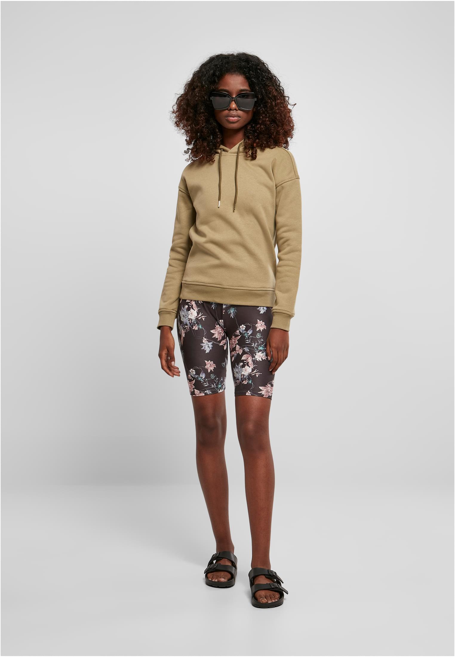 Kvinna fotograferad framifrån i en ljusgrön huvtröja, blommiga cykelbyxor och svarta sandaler, mot en grå bakgrund.