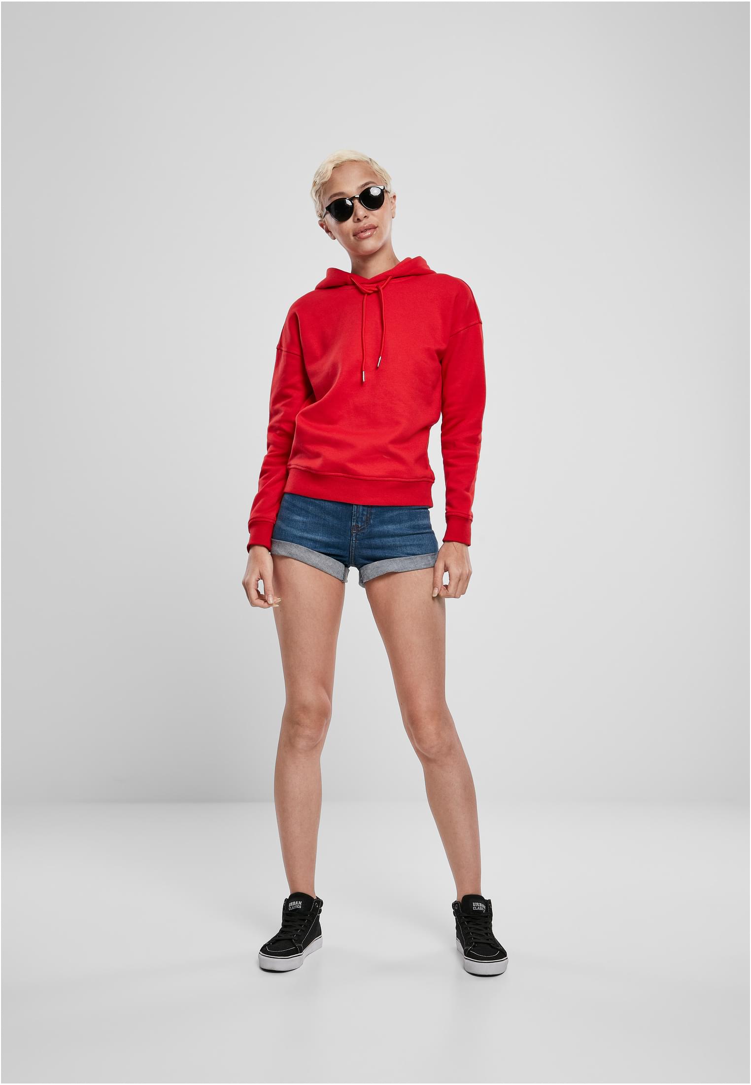 En kvinnlig modell med kort blont hår och solglasögon poserar i en röd huvtröja, jeansshorts och svarta sneakers mot en ljusgrå bakgrund.