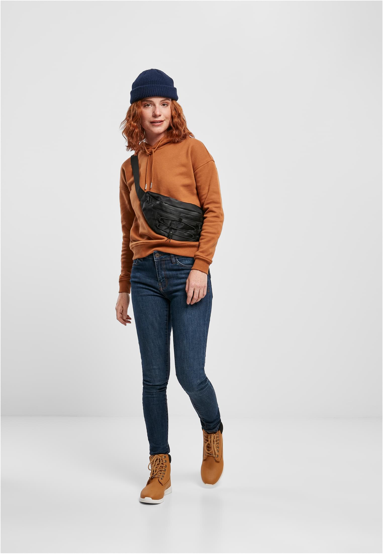 Helkroppsbild av en kvinna som bär en brun huvtröja, blåa jeans, bruna kängor och en blå mössa.