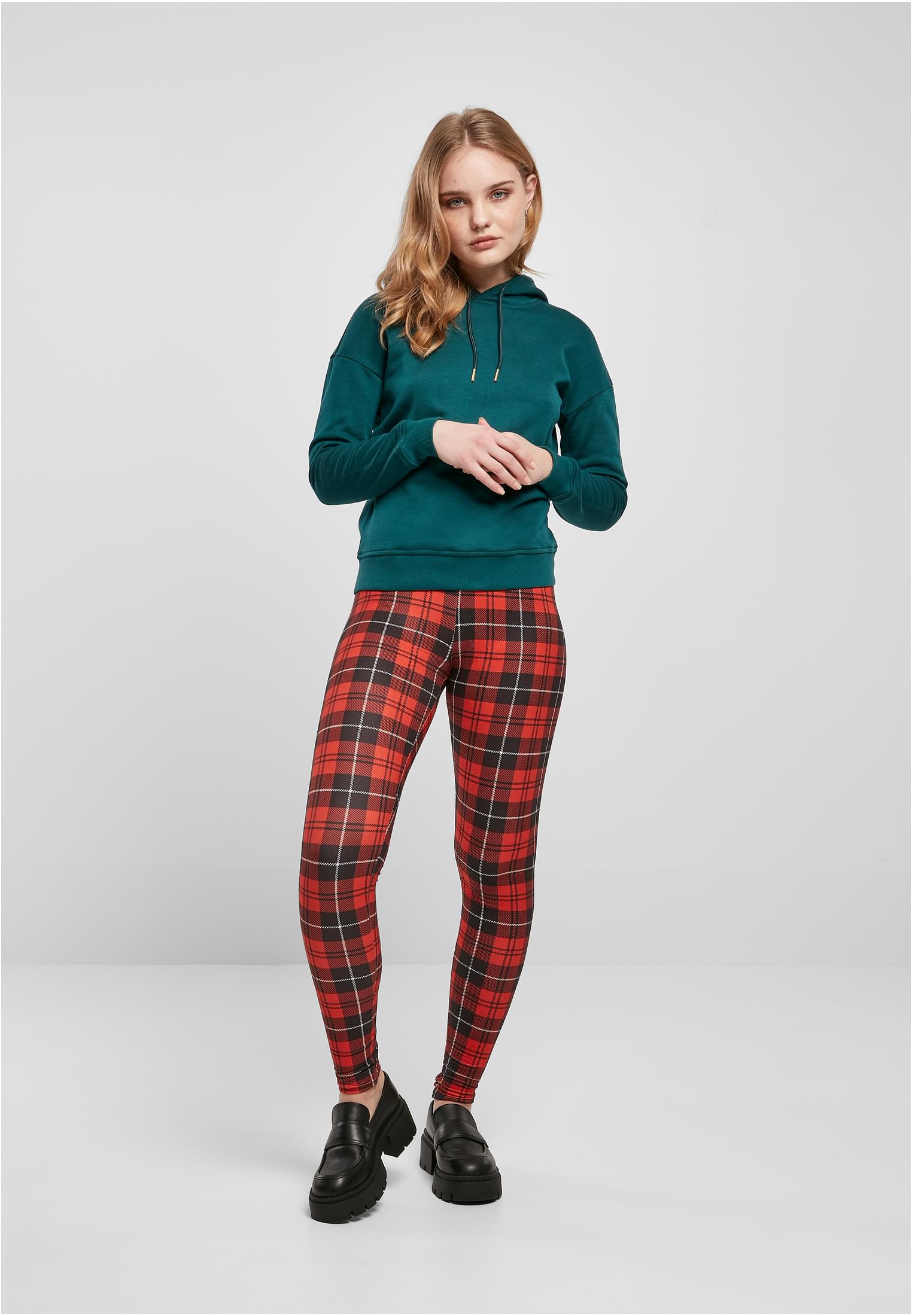 Kvinna som poserar i en mörkgrön luvtröja för damer från Urban Classics, kombinerad med rödrutiga leggings och svarta loafers.