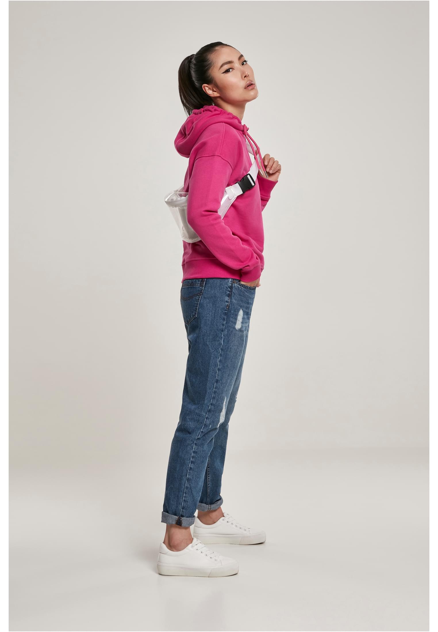 En kvinna som poserar i profil, iförd en klarrosa luvtröja, blå jeans och vita sneakers.