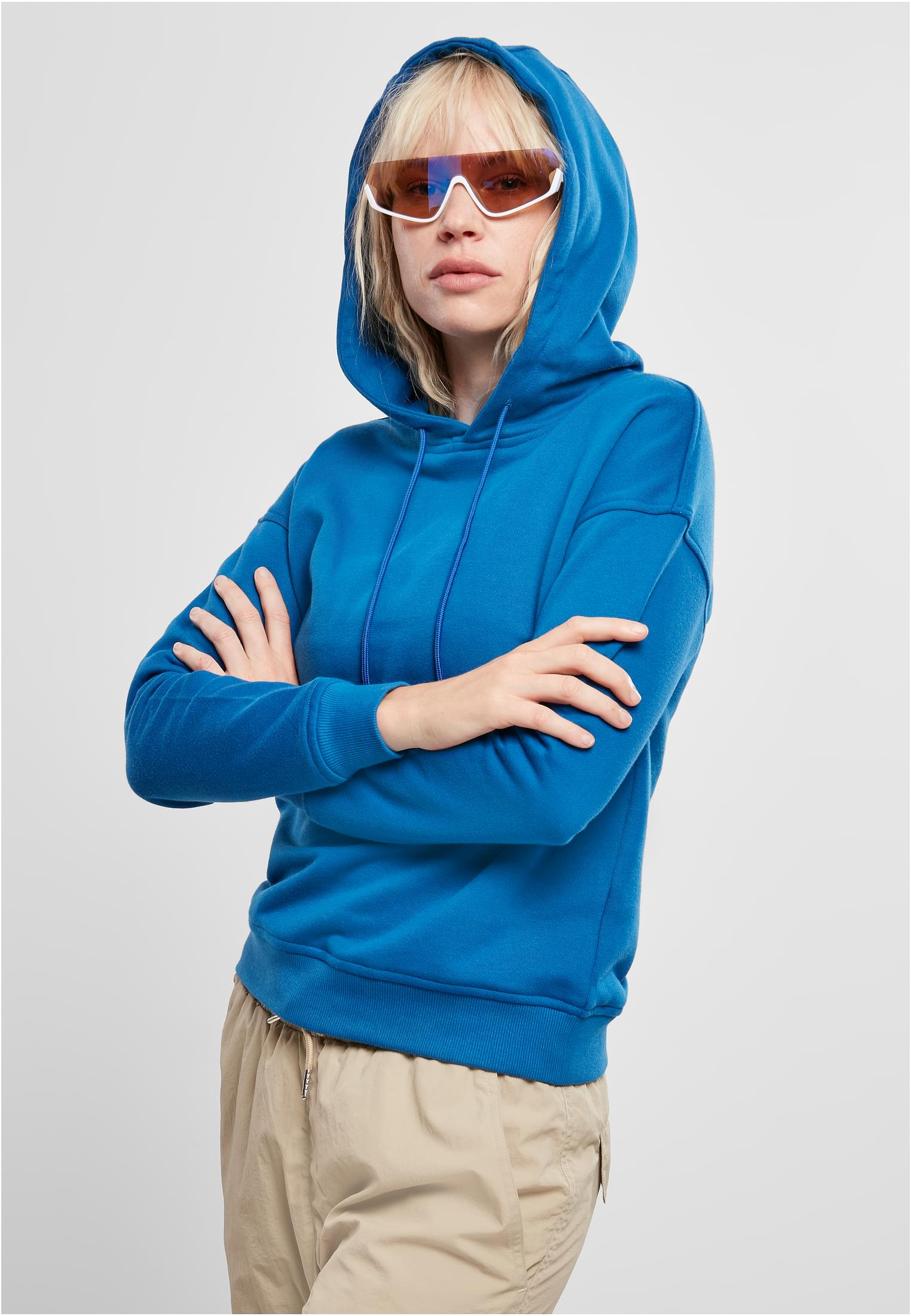 Kvinna med blont hår bär en blå huvtröja med luvan uppe. Hon poserar med armarna i kors mot en vit bakgrund.