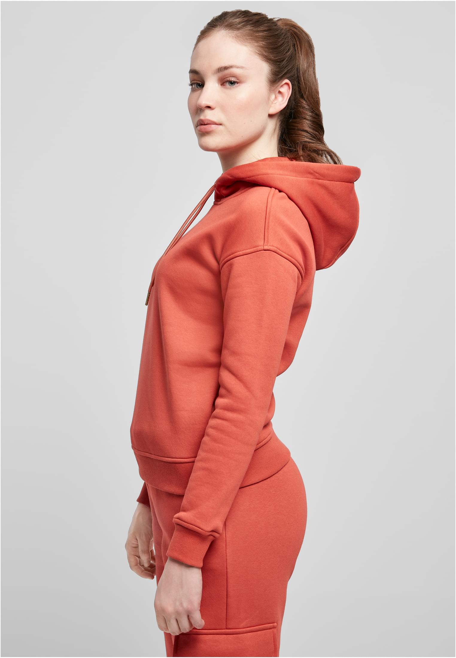 Sidovy av en kvinna som bär en rostfärgad Ladies Hoody från Urban Classics. Hon har håret uppsatt i en hästsvans och tittar över axeln mot kameran.