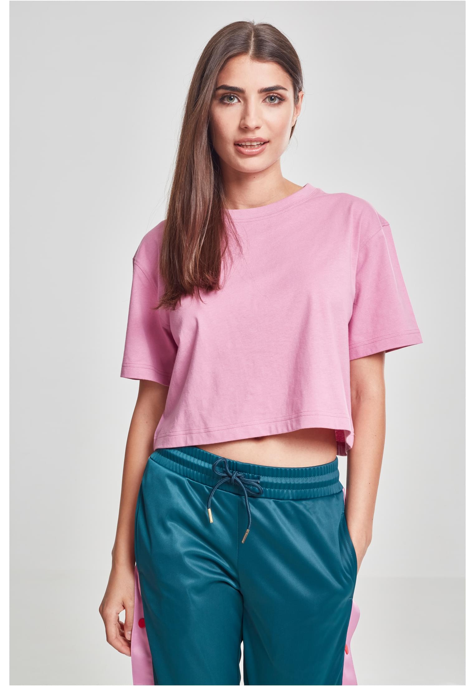 En kvinna som bär en kort oversized rosa t-shirt och turkosa byxor framför en ljusgrå bakgrund.
