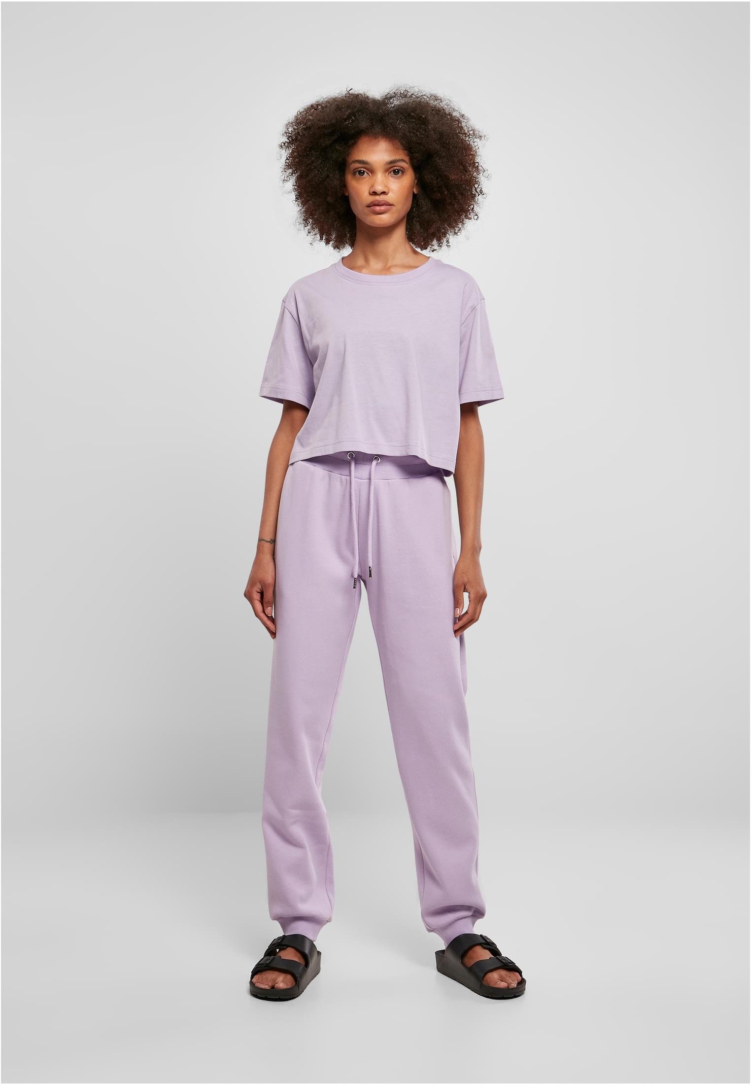 En kvinna som bär en kort, oversized t-shirt i ljuslila med matchande mjukisbyxor och svarta sandaler, stående mot en ljusgrå bakgrund.