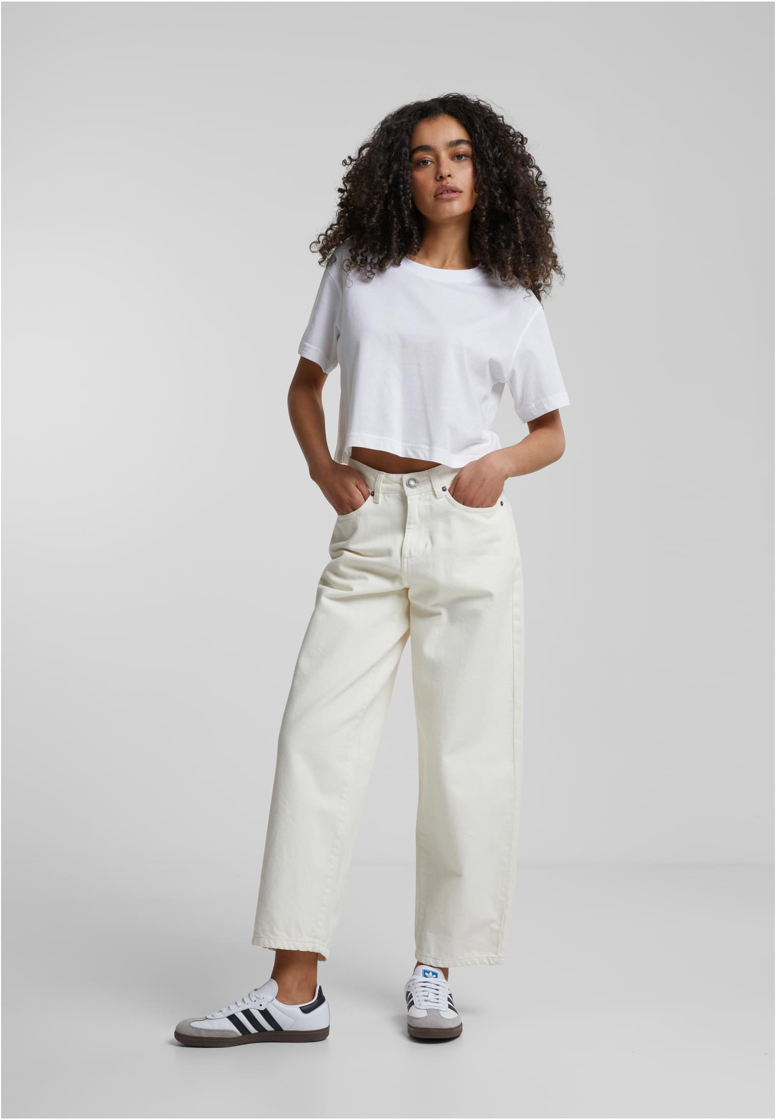 Helbild på en kvinna med lockigt hår som bär en vit, kort oversized t-shirt och ljusa jeans med vida ben.