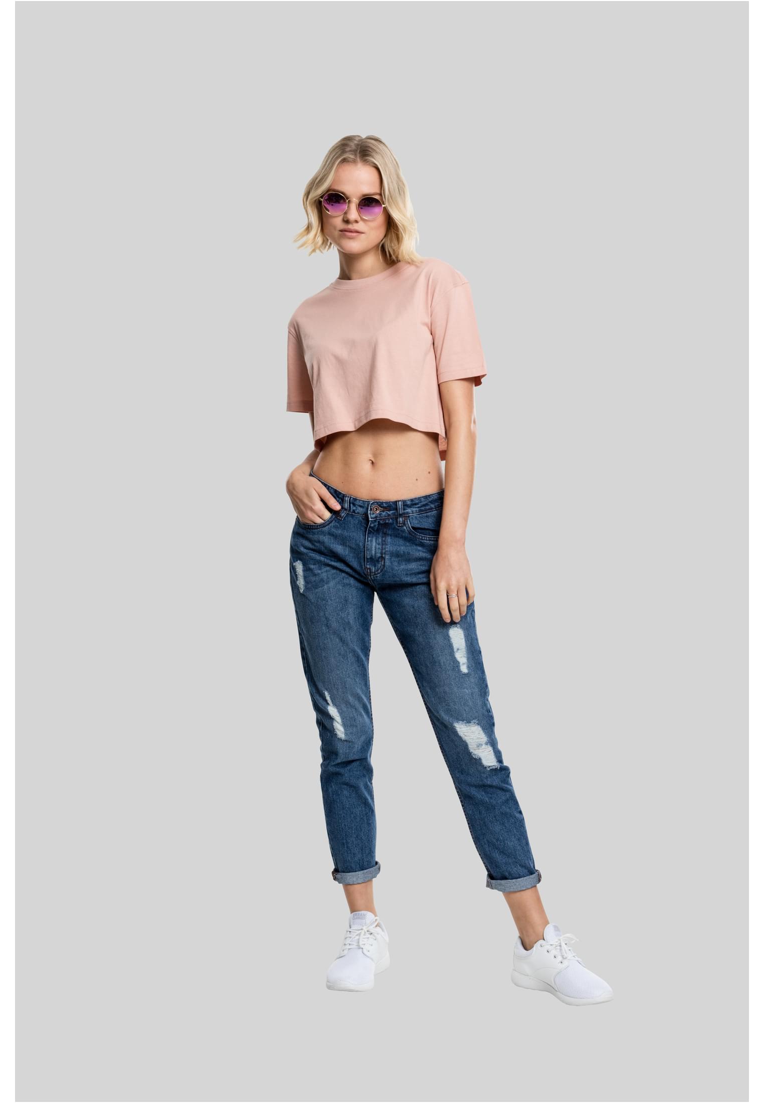 En kvinna poserar i en terrakottafärgad Ladies Short Oversized Tee, slitna jeans och vita sneakers mot en grå bakgrund.