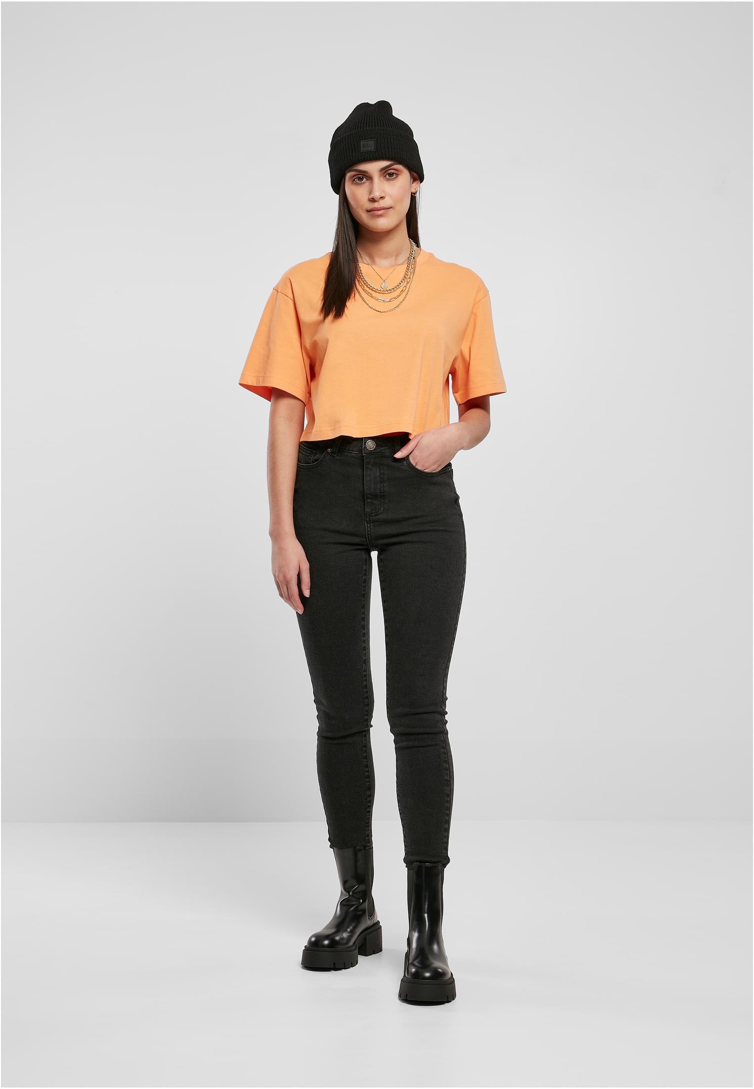 Kvinna som poserar framifrån i en kort, oversized t-shirt i terrakotta, svarta jeans, svarta kängor och en svart mössa.