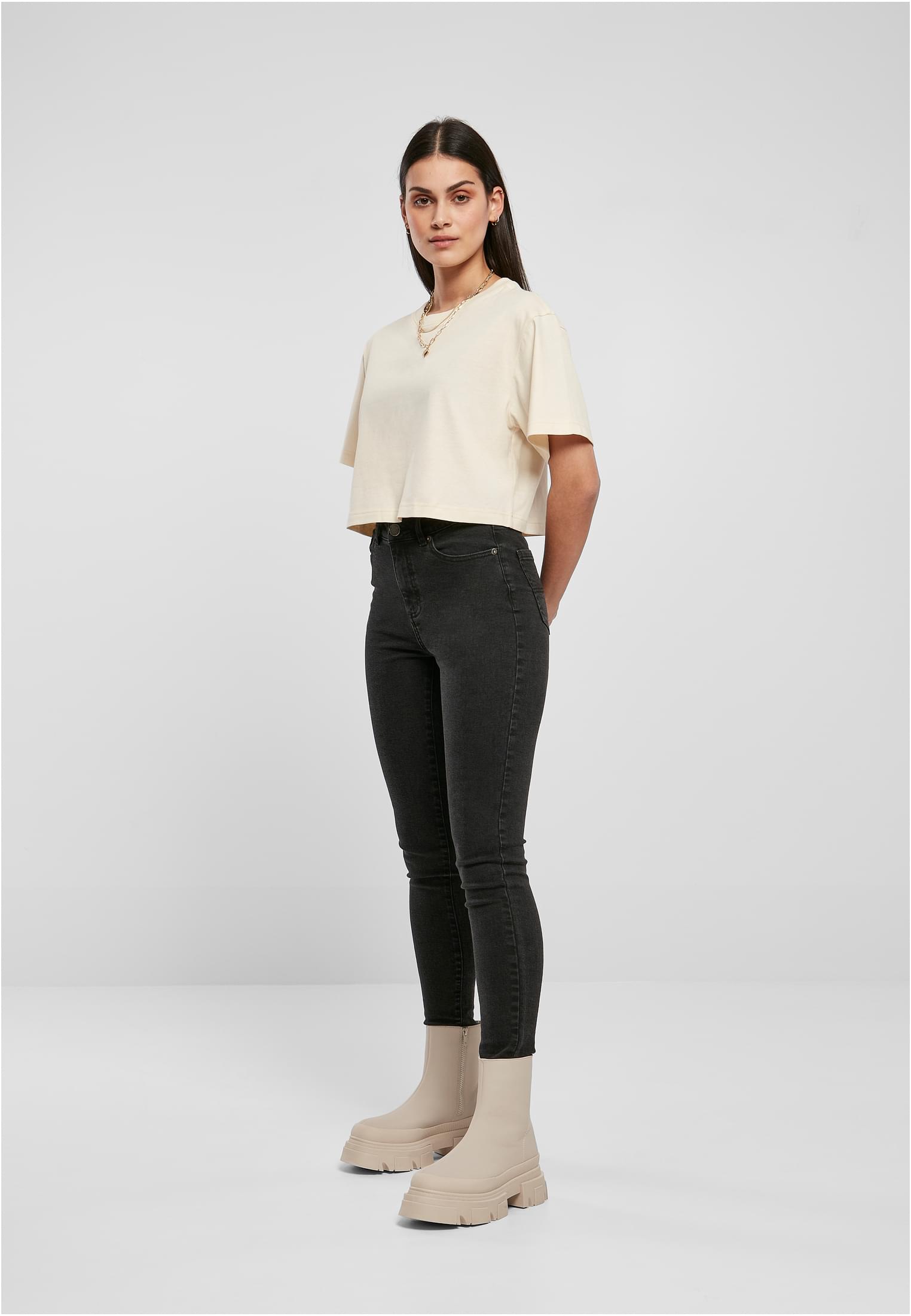 Helbild på en kvinna som poserar i en kort, ljus oversized t-shirt, mörka skinny jeans och ljusa, kraftiga kängor mot en ljusgrå bakgrund.