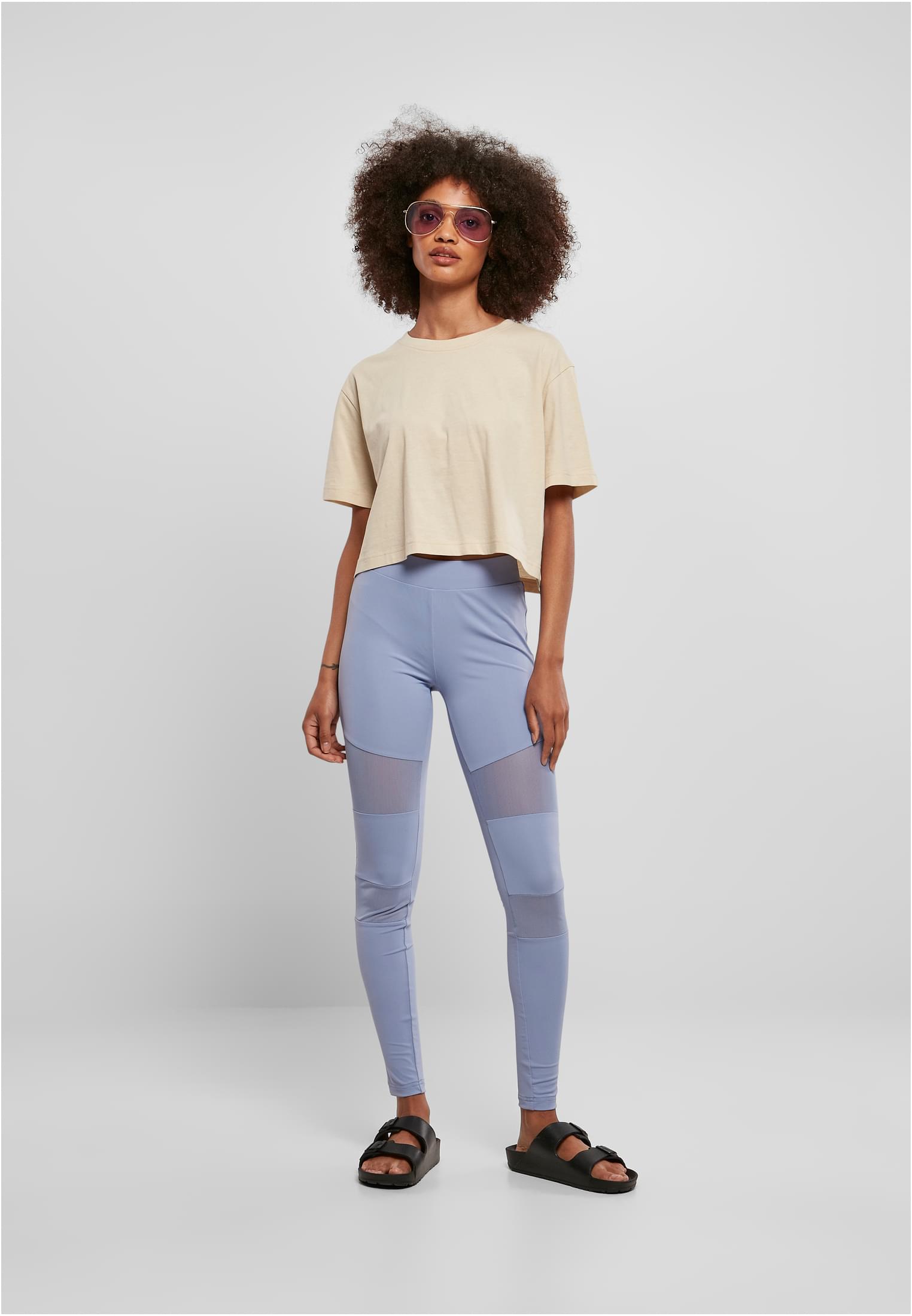 Kvinna sedd framifrån i en kort, oversized terrakottafärgad t-shirt, ljusblå leggings med nätpaneler och svarta sandaler.