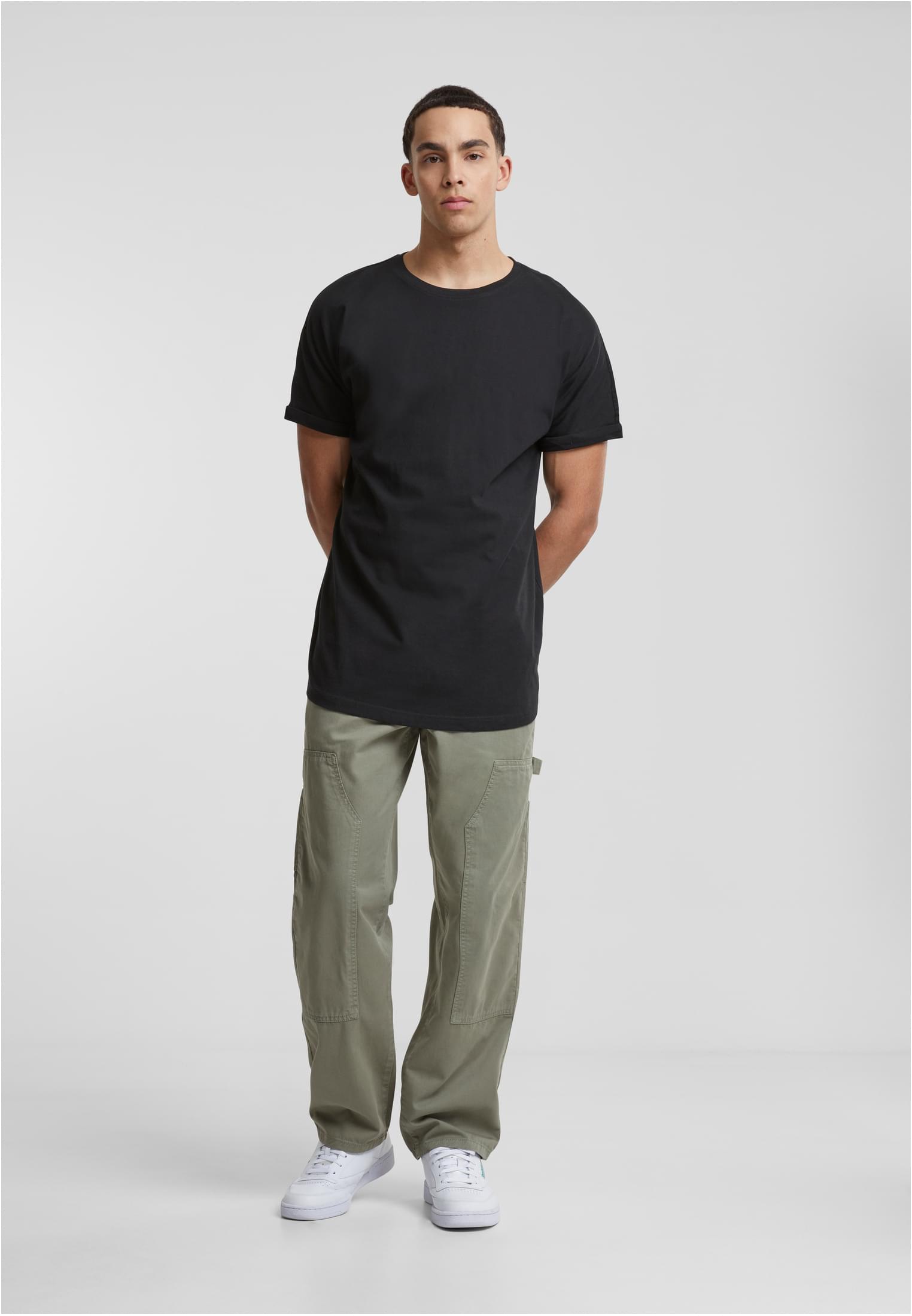 Man i svart t-shirt, olivgröna cargobyxor och vita sneakers. Helkroppsbild tagen framifrån mot en ljusgrå bakgrund.