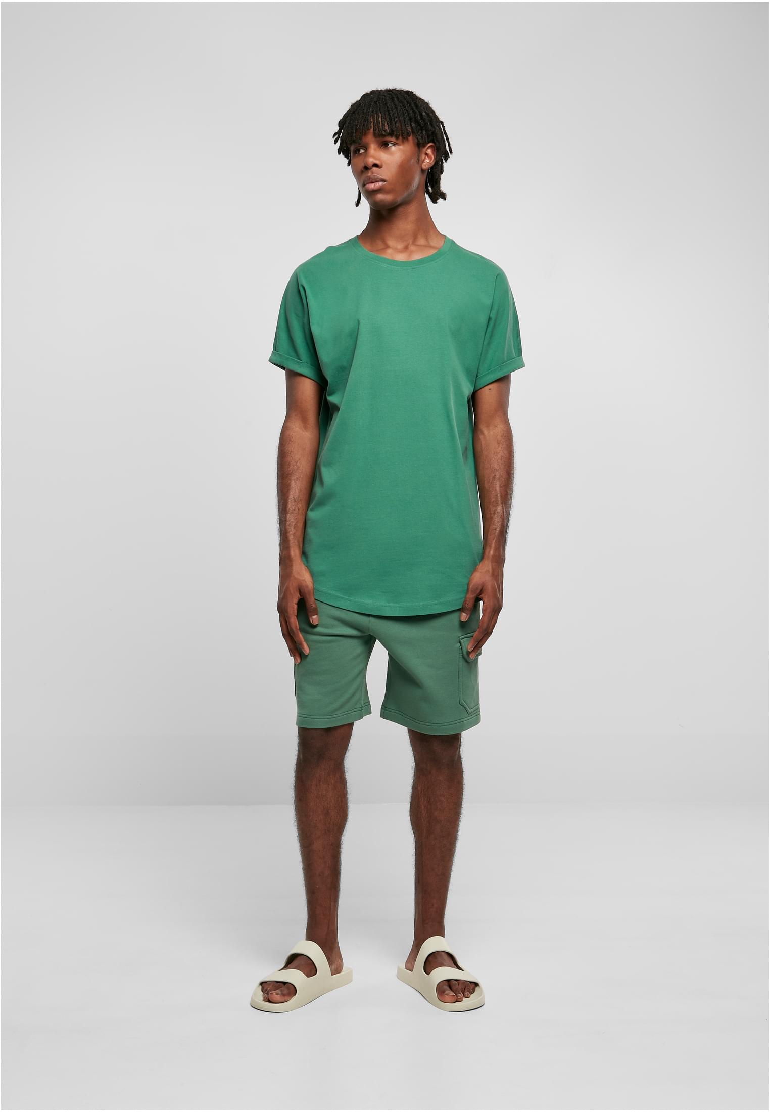 En manlig modell klädd i en grön t-shirt med uppvikta ärmar, gröna shorts och ljusa sandaler.