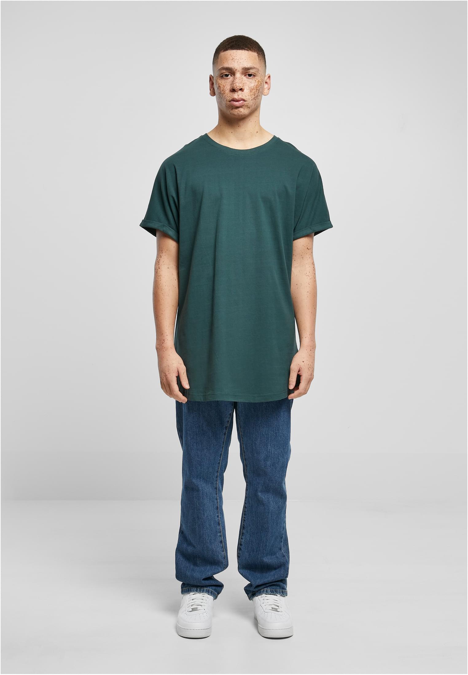 Manlig modell framifrån i en mörkgrön Urban Classics Long Shaped Turnup Tee, blå jeans och vita sneakers.