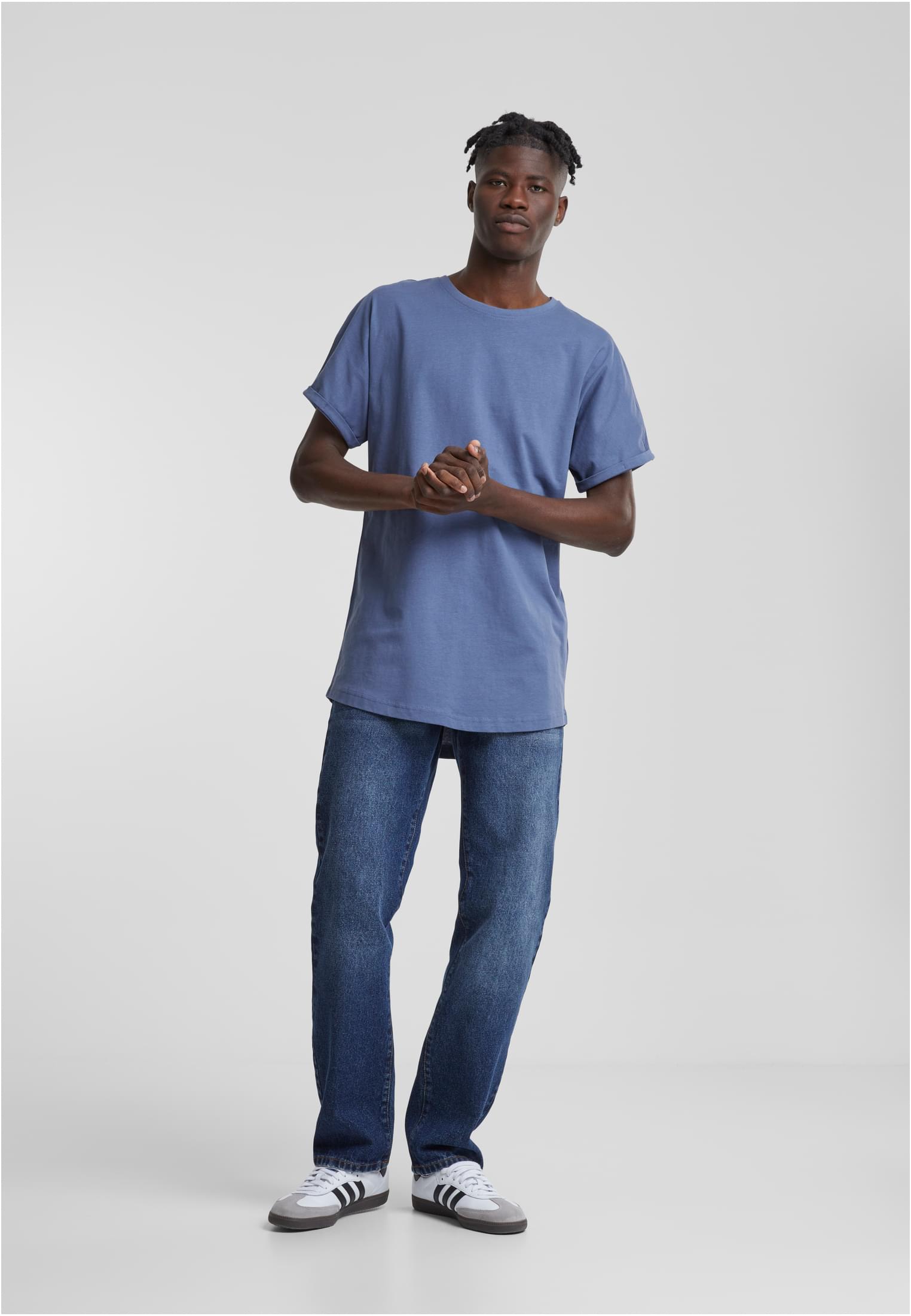 En man i helfigur som bär en blå t-shirt med uppvikta ärmar, blå jeans och vita sneakers, stående mot en ljusgrå bakgrund.