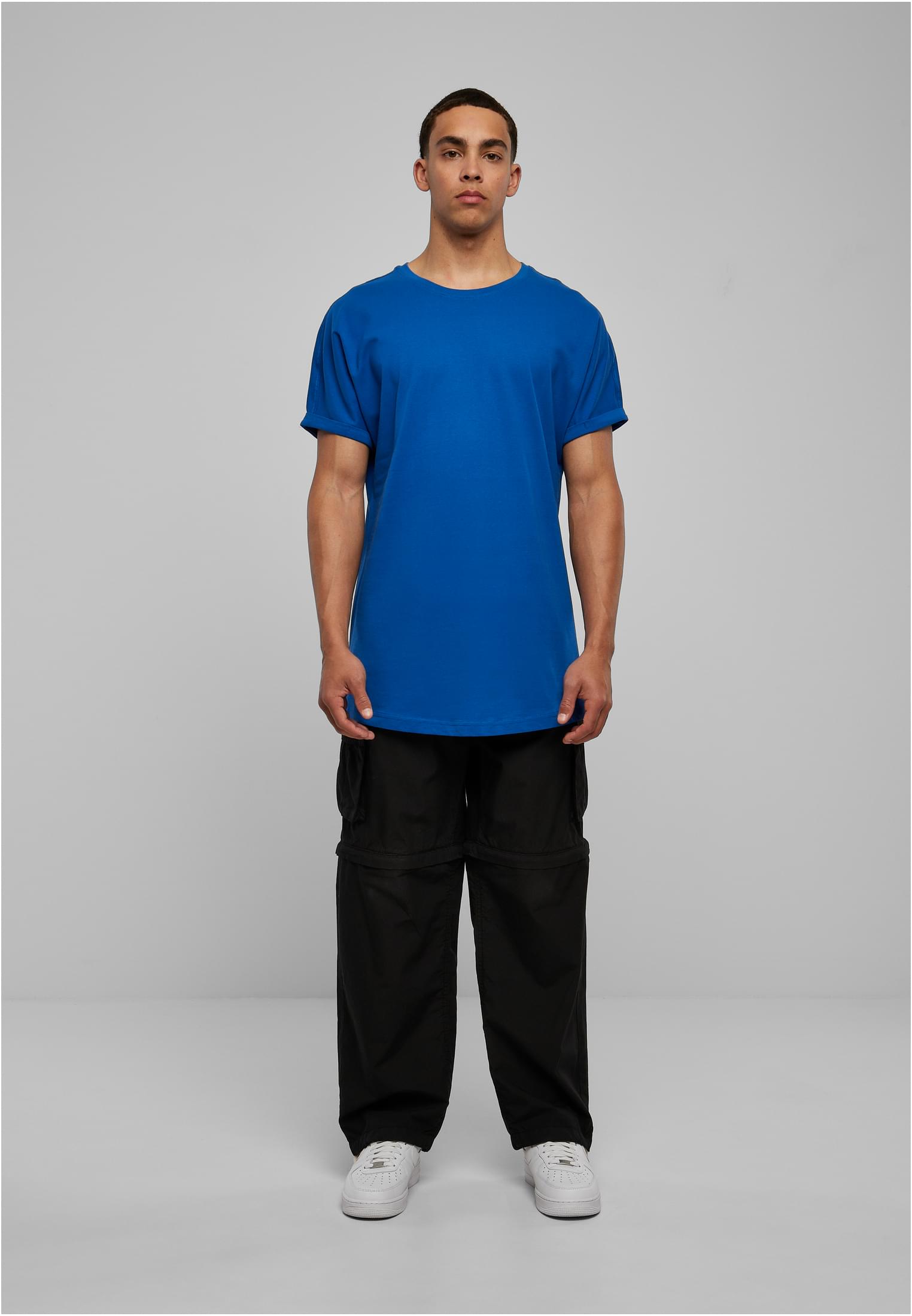 En manlig modell poserar i en blå Long Shaped Turnup Tee med svarta byxor och vita skor.