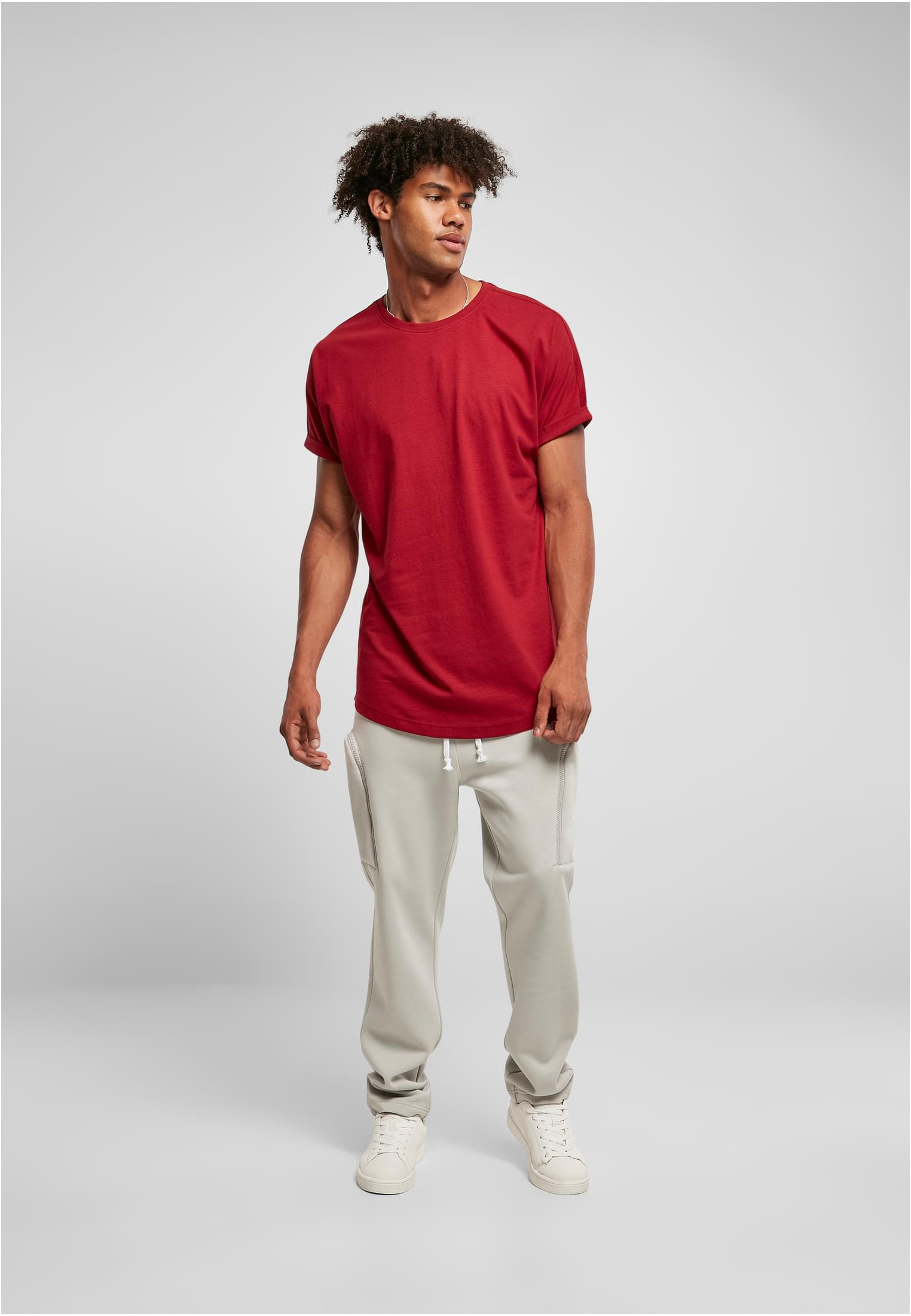 En manlig modell poserar i en terrakottafärgad Long Shaped Turnup Tee, ljusgrå mjukisbyxor och vita sneakers framför en ljusgrå bakgrund.