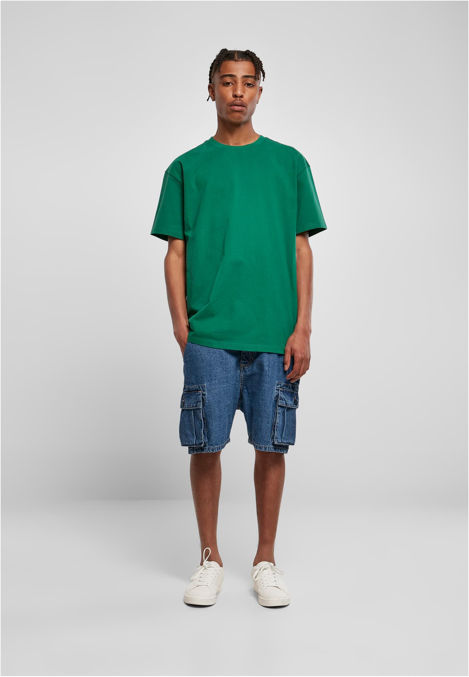 Manlig modell som bär en grön oversized t-shirt, blåa cargoshorts i denim och vita sneakers, stående mot en ljusgrå bakgrund.