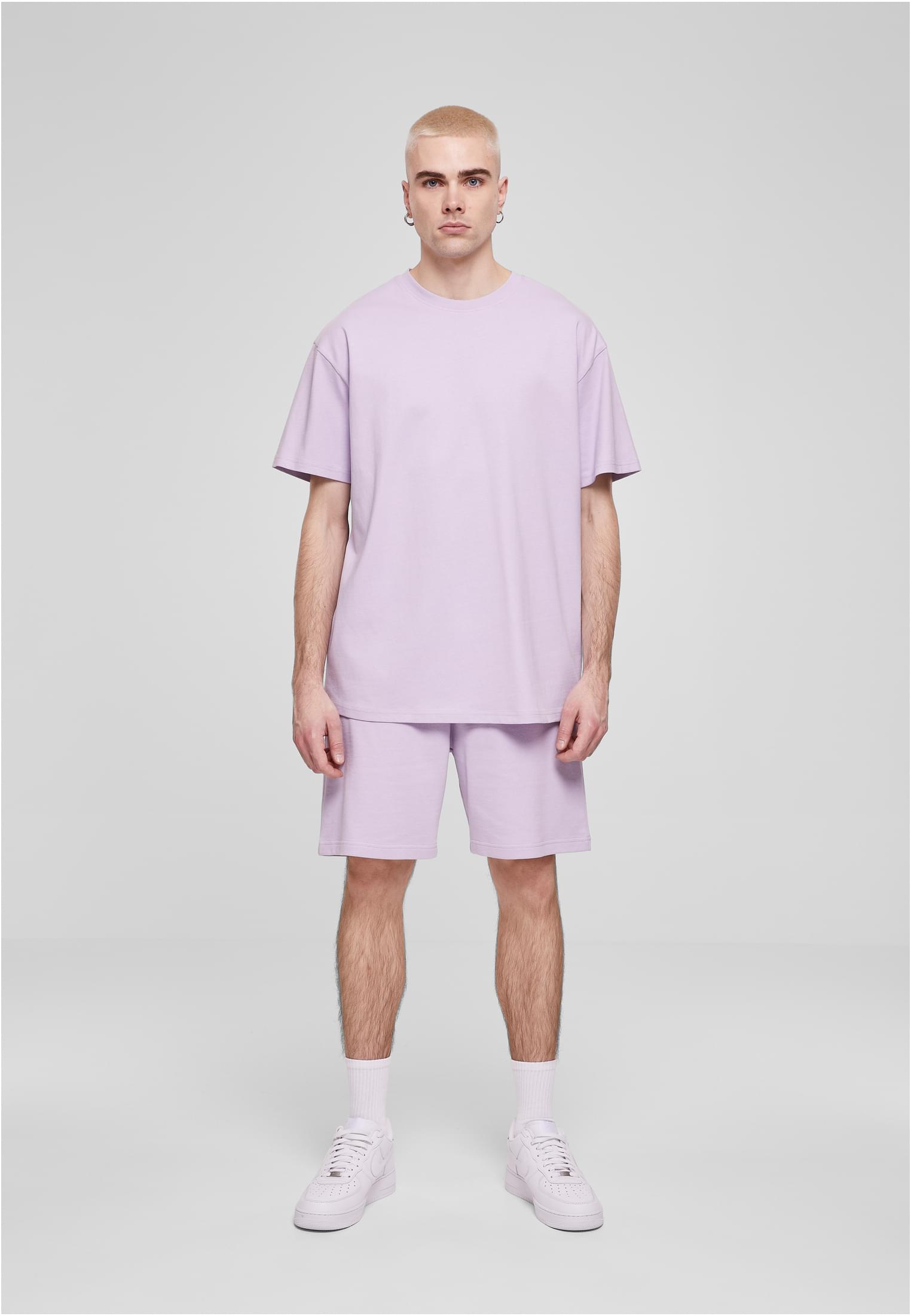 En manlig modell med blonderat hår bär en ljuslila oversized t-shirt och matchande shorts, med vita strumpor och vita sneakers, framför en ljusgrå bakgrund.
