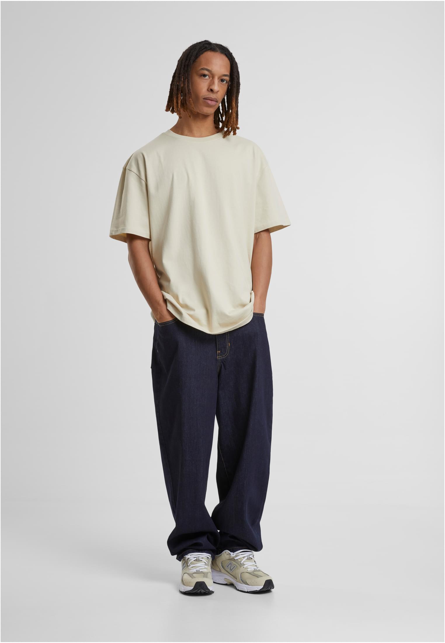 En manlig modell poserar i en krämvit oversized t-shirt, mörkblå vida jeans och ljusa sneakers mot en vit bakgrund.