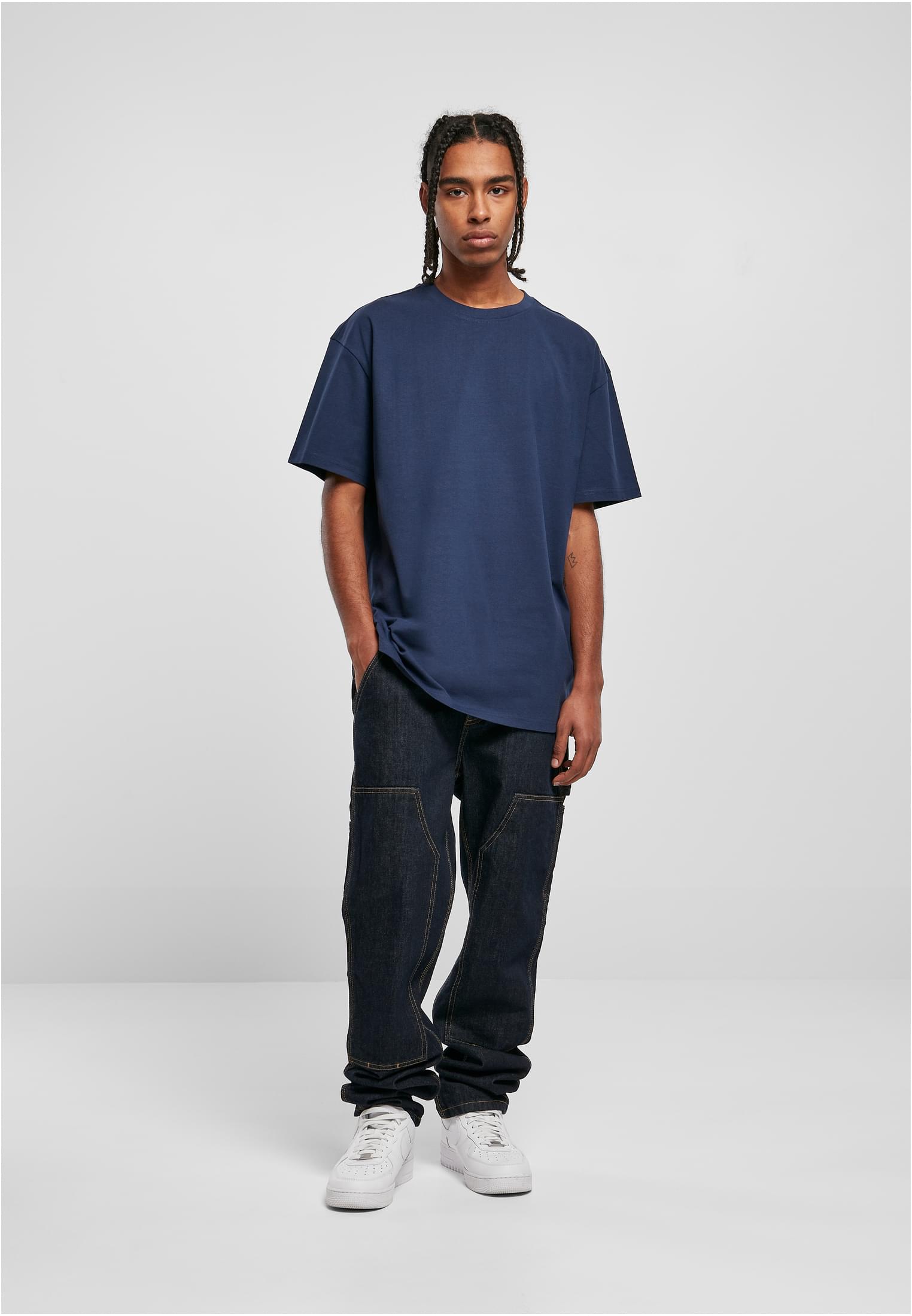 En manlig modell poserar i en mörkblå oversized t-shirt, mörka jeans och vita sneakers mot en ljusgrå bakgrund.