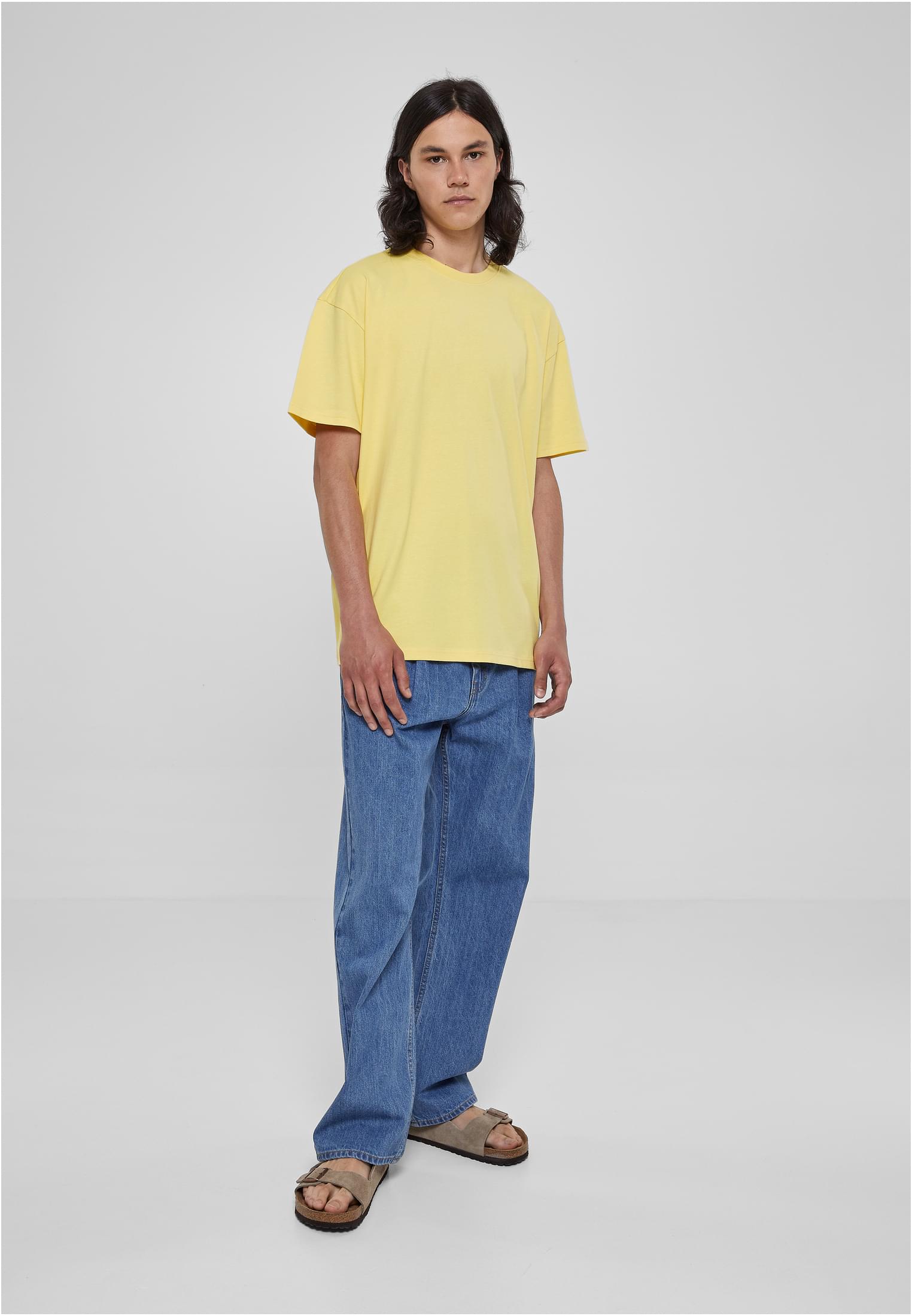 En manlig modell som bär en gul Heavy Oversized Tee med vida blåa jeans och bruna sandaler framför en ljusgrå bakgrund.