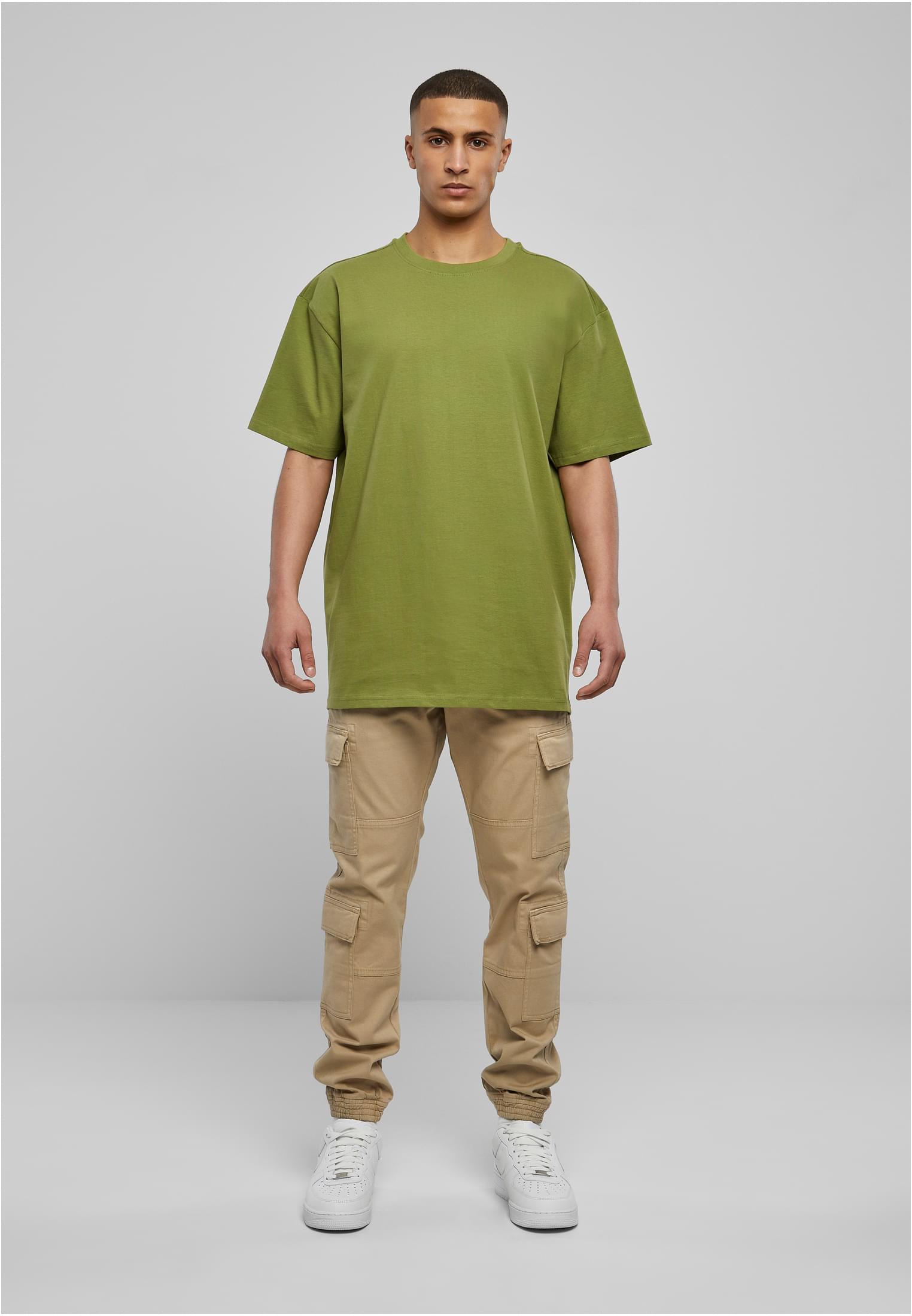 Man som bär en grön Heavy Oversized Tee, beiga cargobyxor och vita sneakers mot en ljusgrå bakgrund.