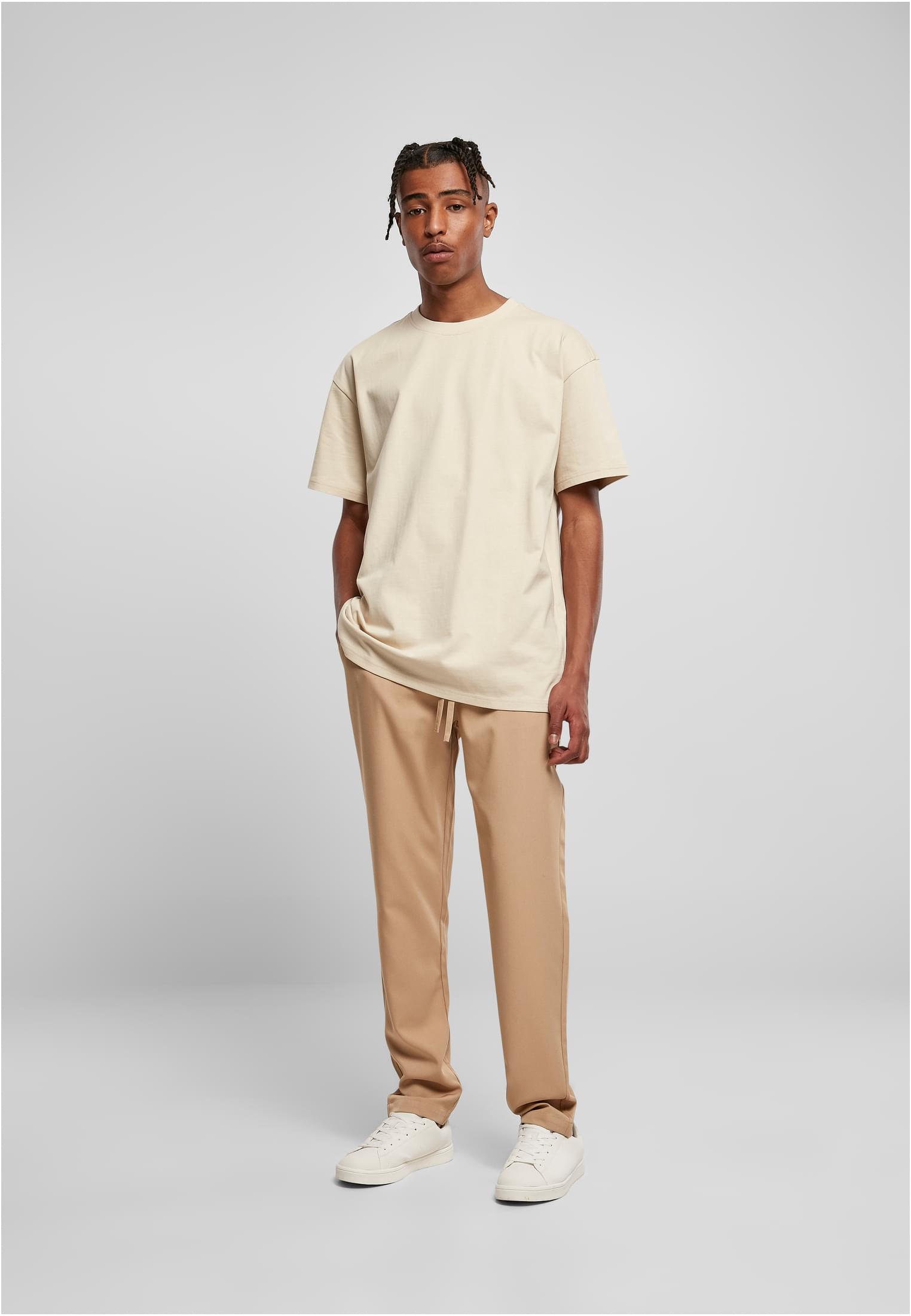 En manlig modell som poserar i en ljus oversized t-shirt, beiga byxor och vita sneakers mot en enfärgad grå bakgrund.