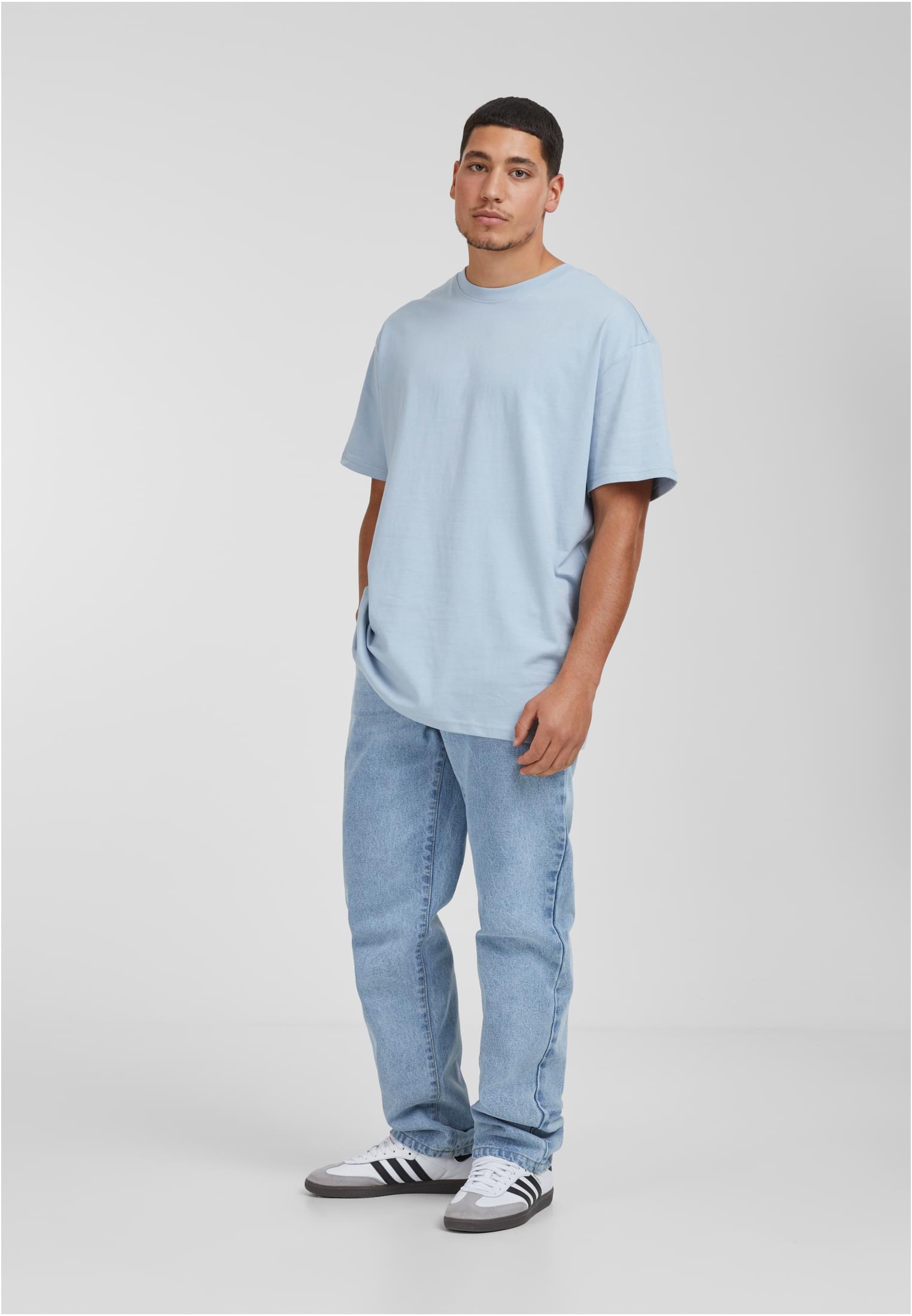 En man poserar i en ljusblå oversized t-shirt, ljusa jeans och vita sneakers med svarta ränder mot en ljusgrå bakgrund.