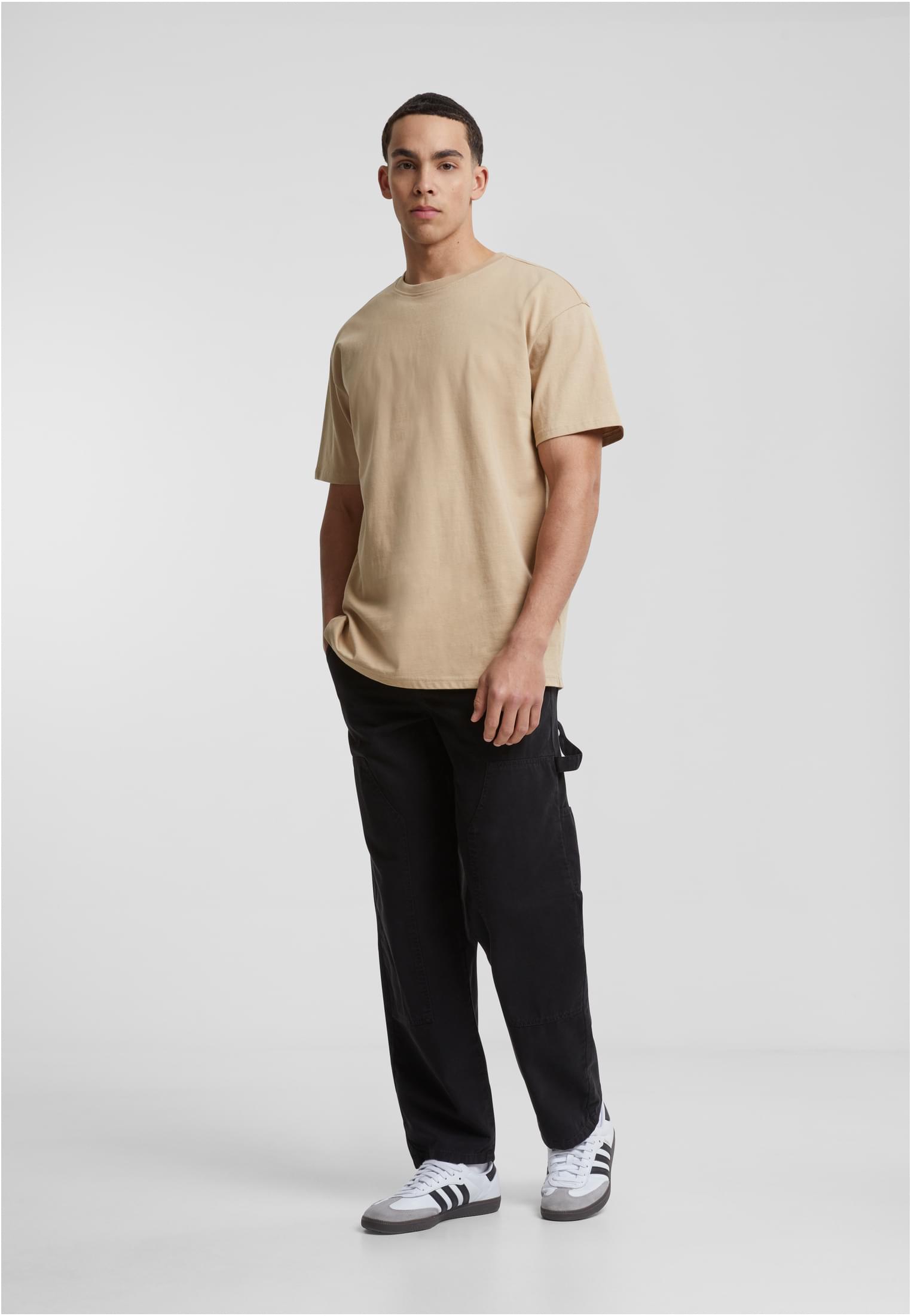 En manlig modell som bär en beige Heavy Oversized Tee från Urban Classics, med svarta byxor och sneakers.