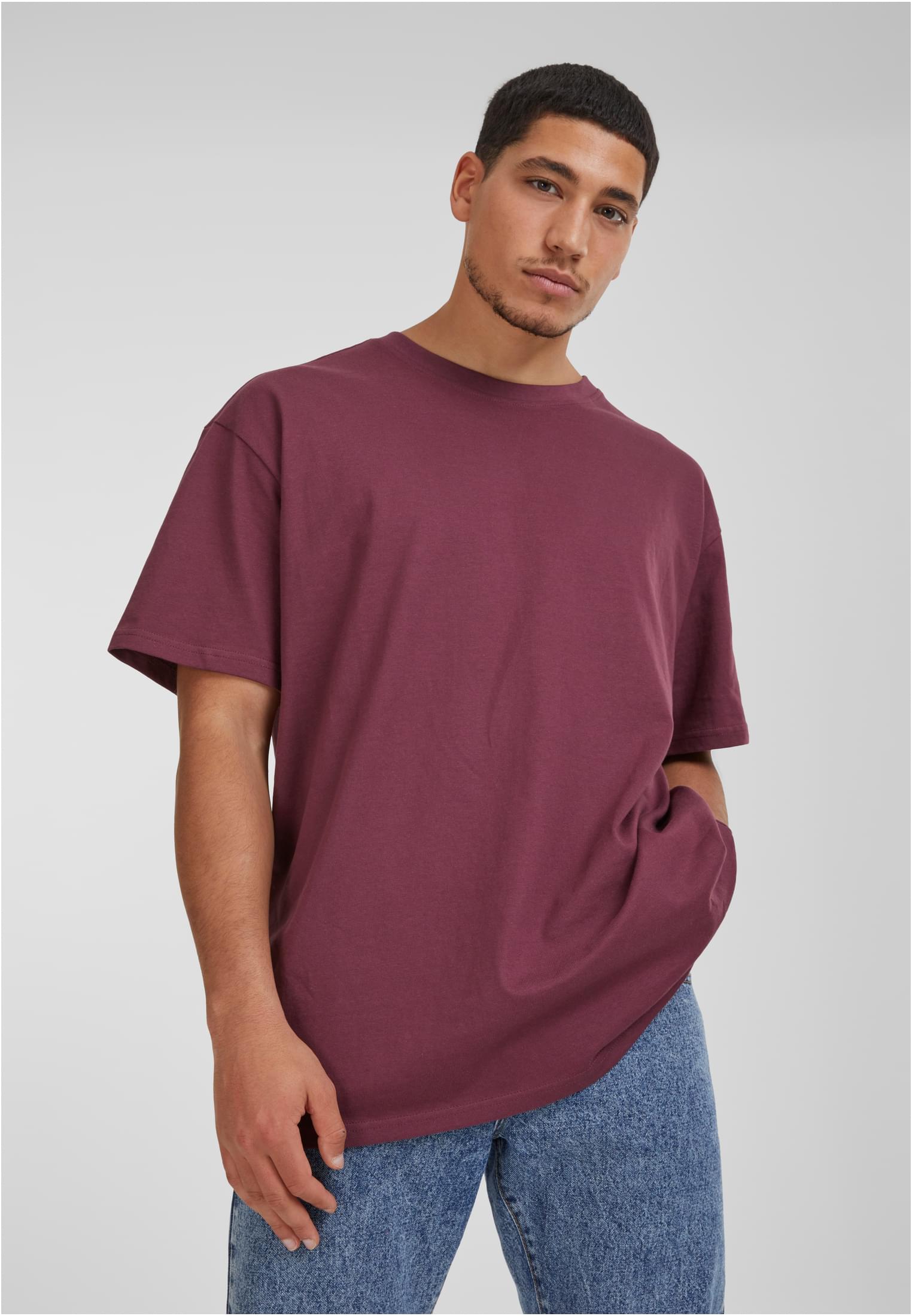 Manlig modell som bär en vinröd oversized t-shirt och blå jeans, fotograferad framifrån mot en grå bakgrund.
