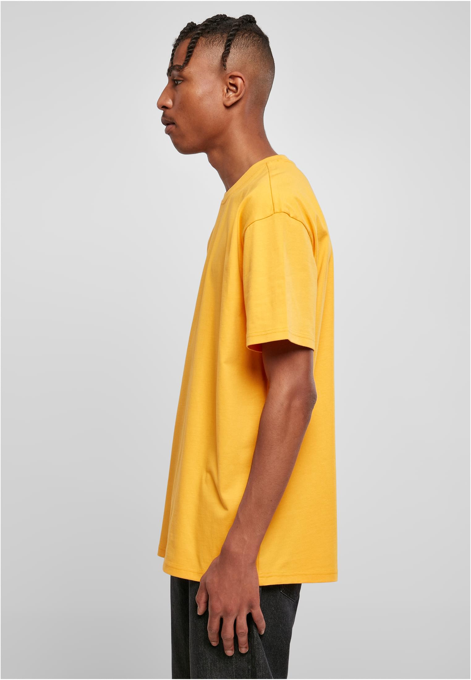 En man sedd från sidan, klädd i en gul oversized t-shirt och mörka byxor mot en ljusgrå bakgrund.