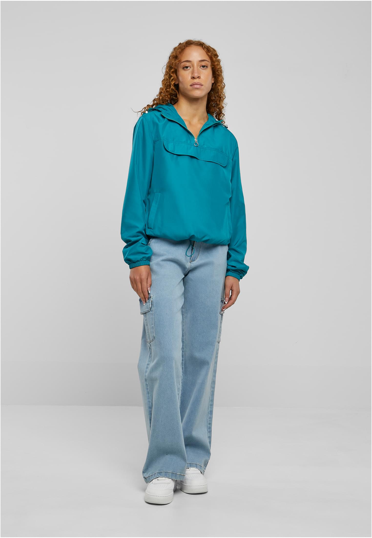 Frontvy av en kvinna som bär den turkosa UC Ladies Basic Pull Over Jacket med ljusblåa vida jeans och vita sneakers.