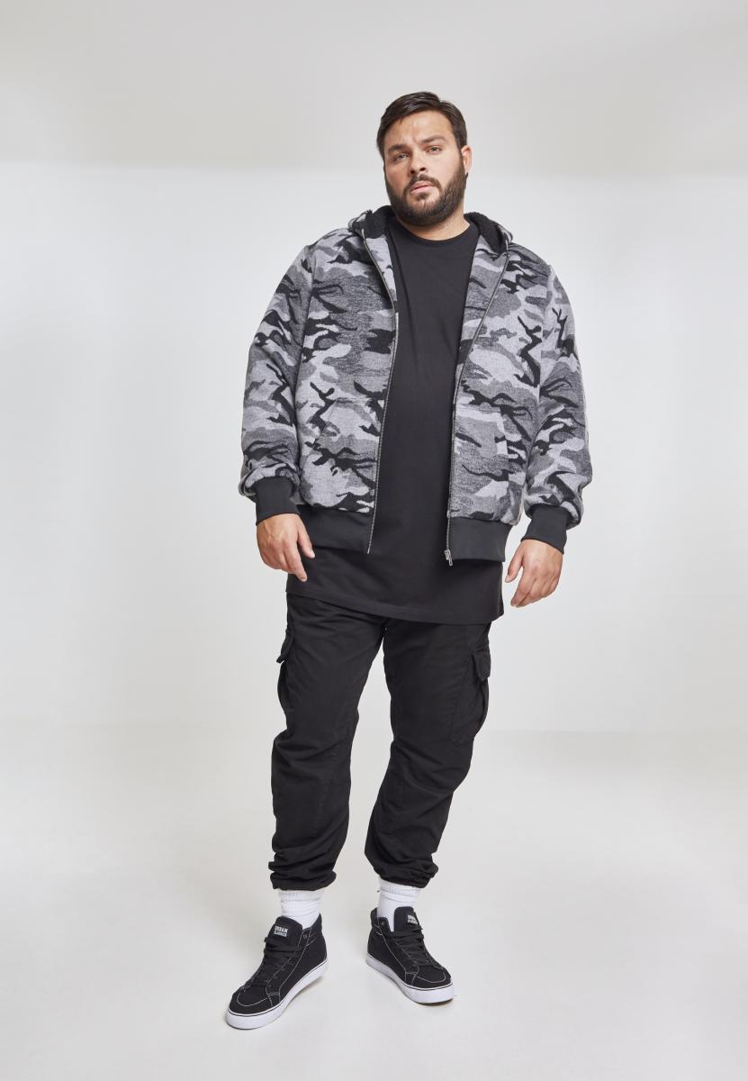 En man bär en öppen UC Camo Zip Jacket med grått kamouflagemönster, en svart t-shirt och svarta byxor mot en vit bakgrund.