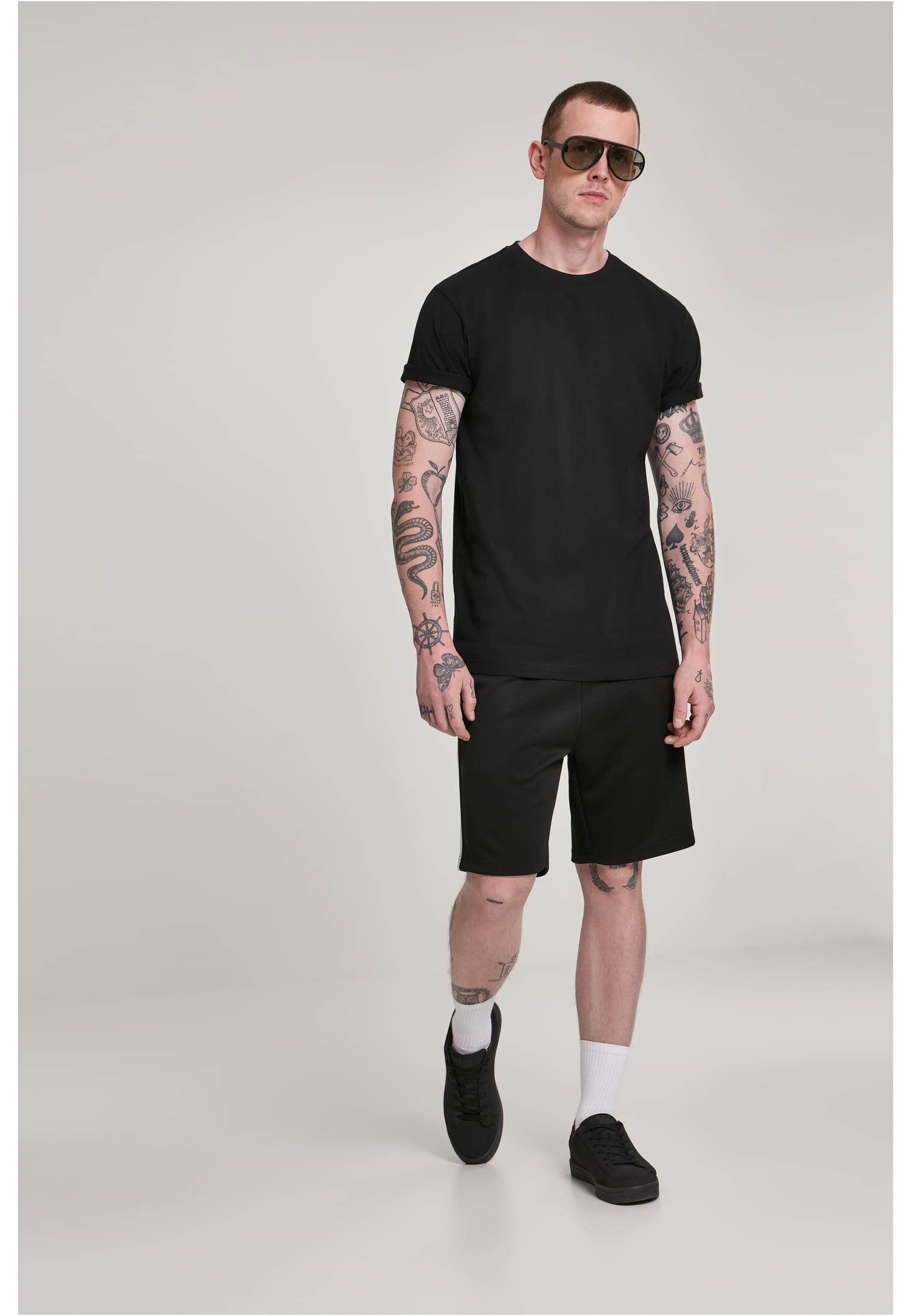 En tatuerad man i solglasögon poserar i en svart t-shirt och svarta shorts.