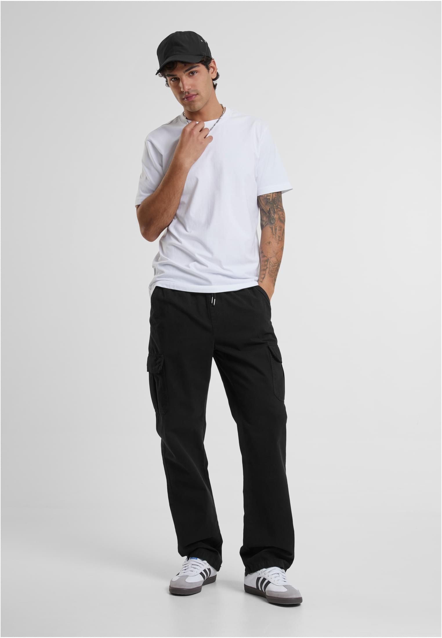 Helbild på en man som bär en vit t-shirt, svarta cargobyxor och en svart keps.