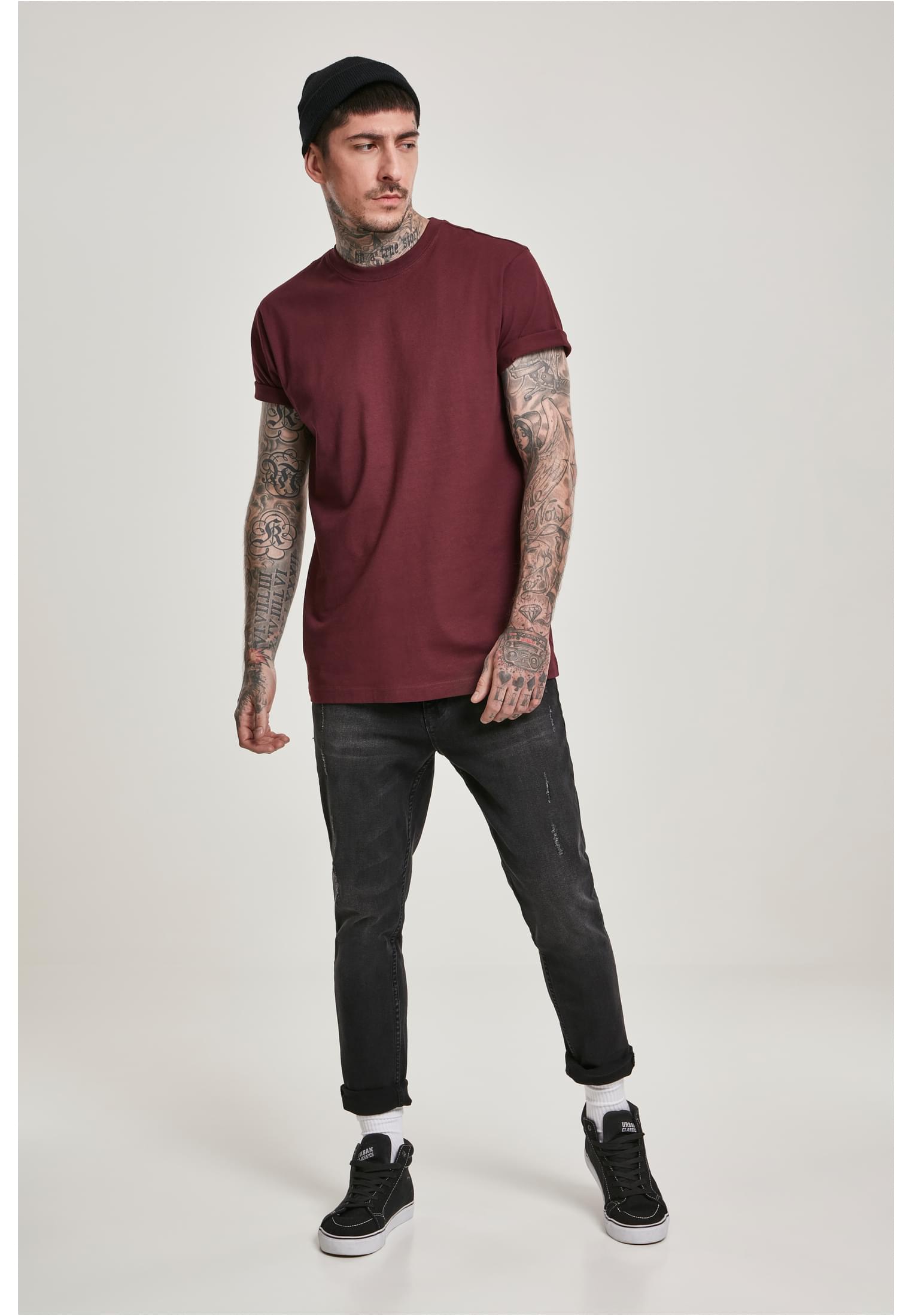 En man med tatueringar poserar i en vinröd Basic Tee, svarta jeans och en mössa.